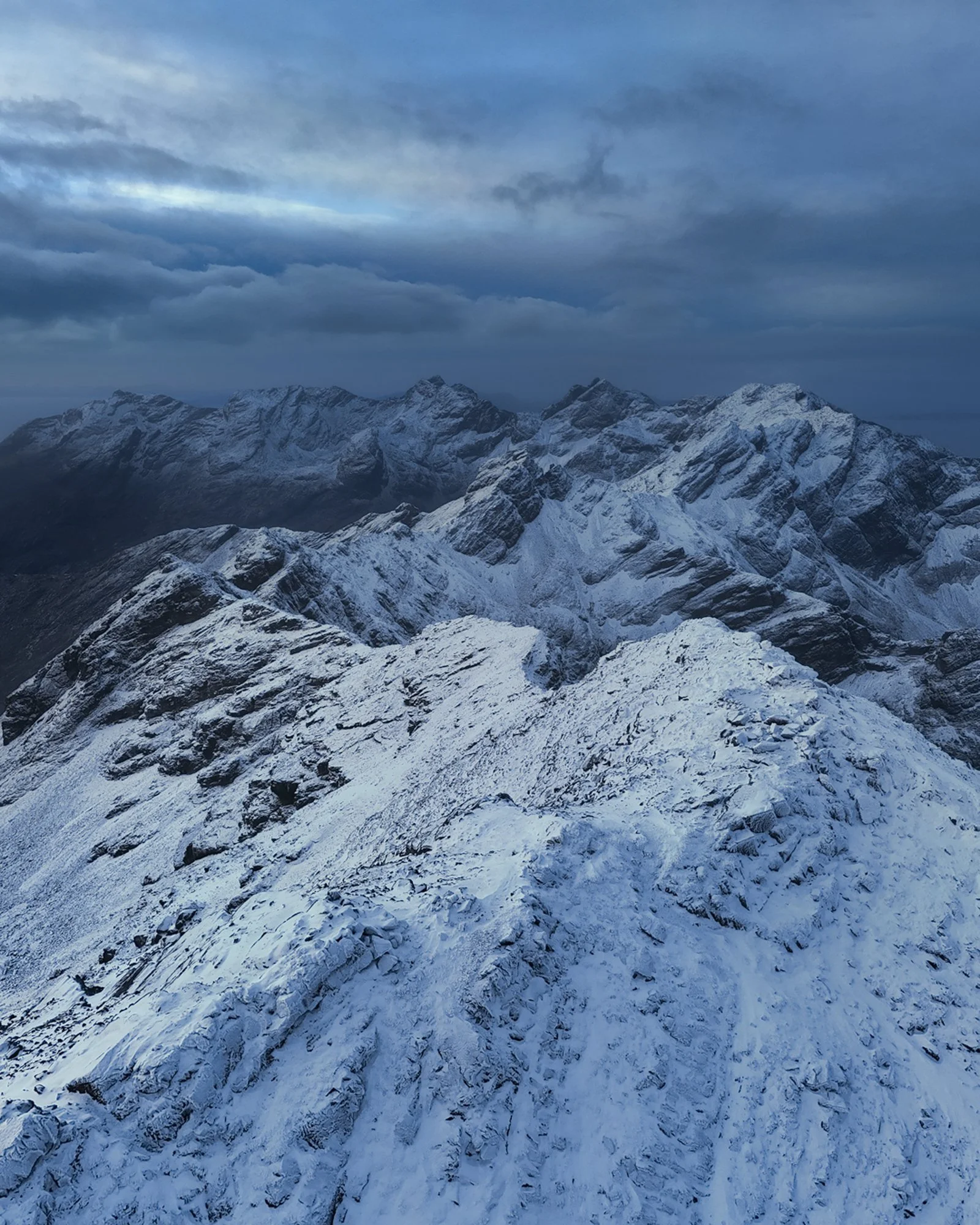 A sweeping aerial view of the jagged Cuillin Ridge on the Isle of Skye, blanketed in fresh snow under an overcast, brooding sky. The rugged peaks rise sharply, their intricate, craggy surfaces highlighted by soft alpenglow that subtly illuminates the