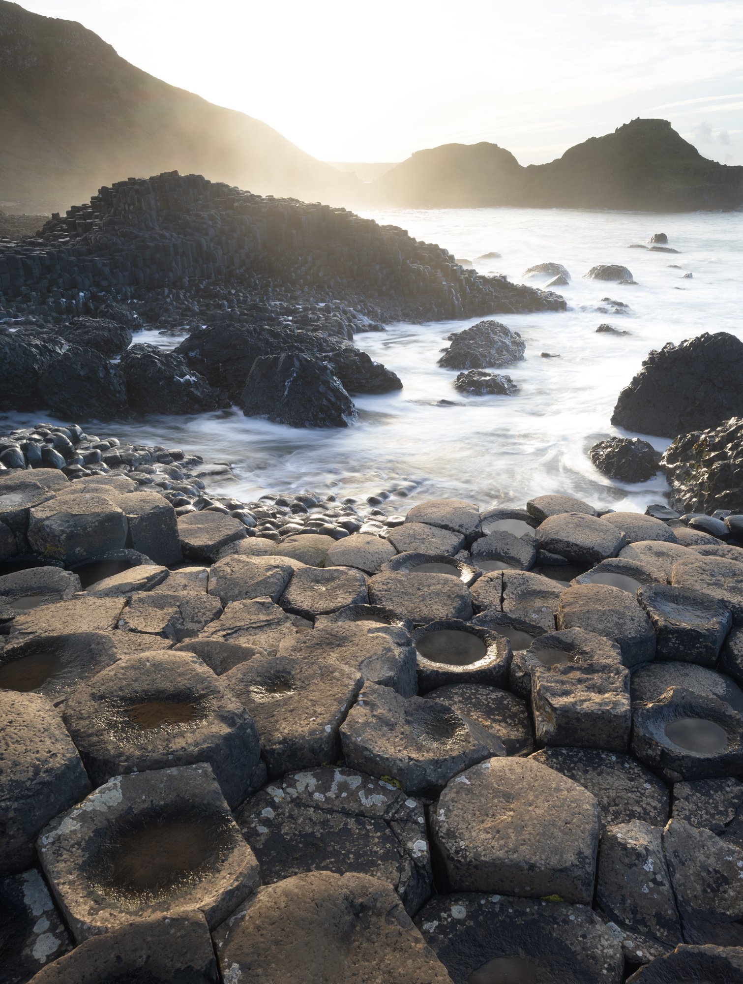 Golden light floods the Giant’s Causeway, illuminating the iconic hexagonal basalt columns as powerful waves crash against the shore. The foam and mist from the ocean add a soft atmospheric touch to the rugged landscape. The unique perspective highli