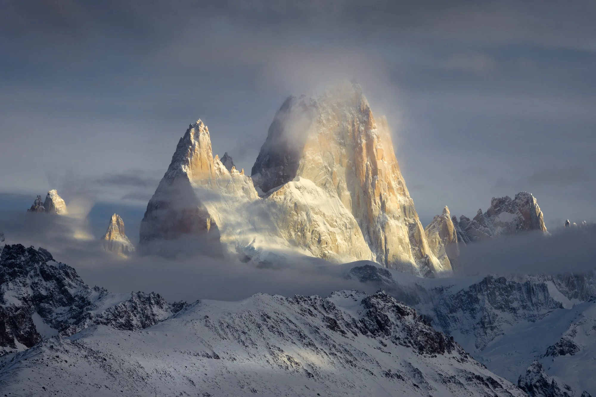 Golden light gently illuminates the rugged, snow-covered peaks of Mount Fitz Roy during sunrise, while mist and low clouds drift across the landscape, adding depth and atmosphere. The telephoto perspective enhances the grandeur of the mountain, highl