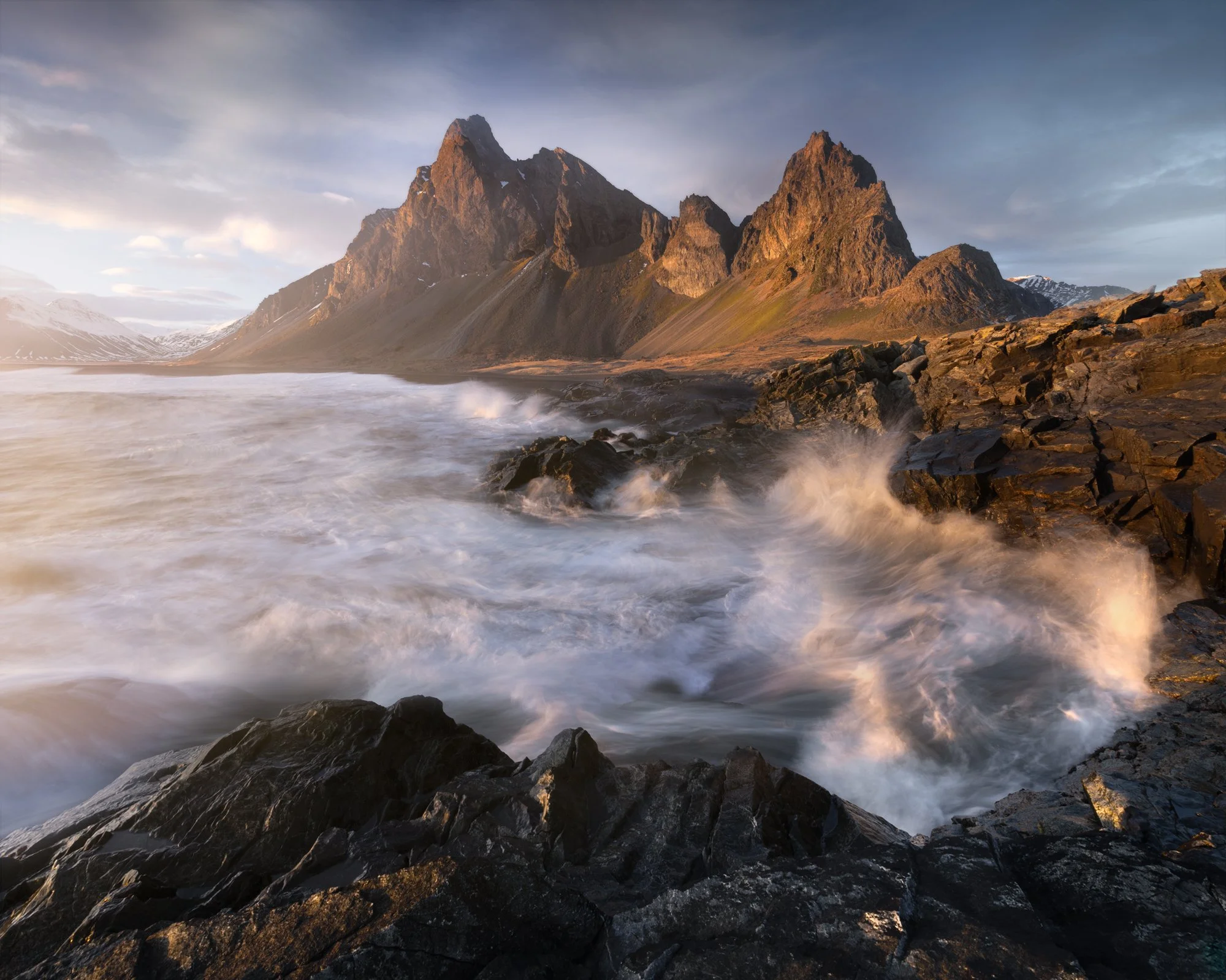 A dramatic coastal scene at Estrahorn, Iceland, with towering mountains catching the golden light of sunset. The powerful waves crash against the jagged black rocks, sending plumes of sea spray into the air. The dynamic contrast between the serene li