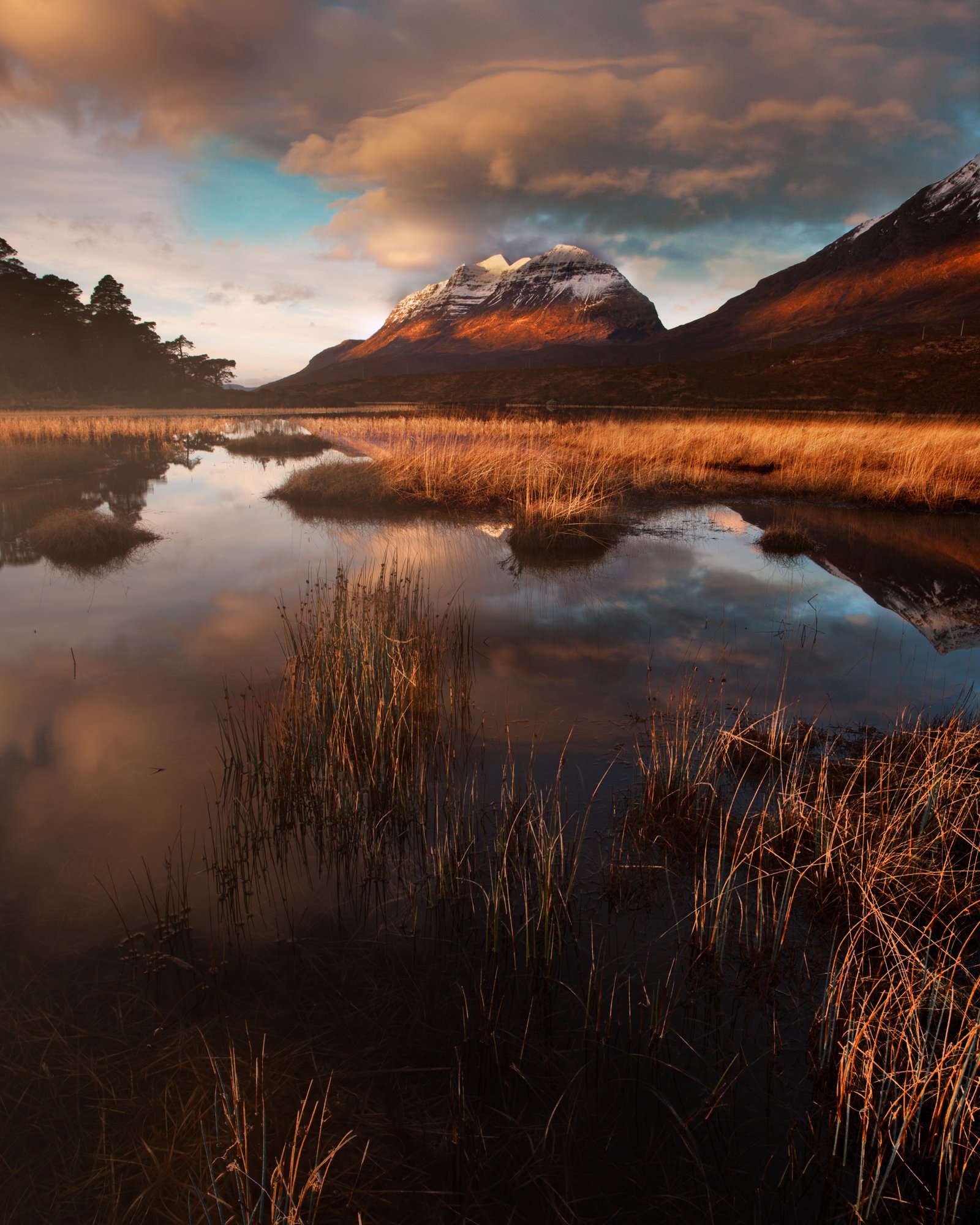 A serene sunrise scene at Torridon’s Loch Clair, bathed in warm, golden light. Snow-dusted mountains stand majestically in the background, reflecting vividly on the calm waters of the loch. The foreground grasses add texture and depth, gently illumin