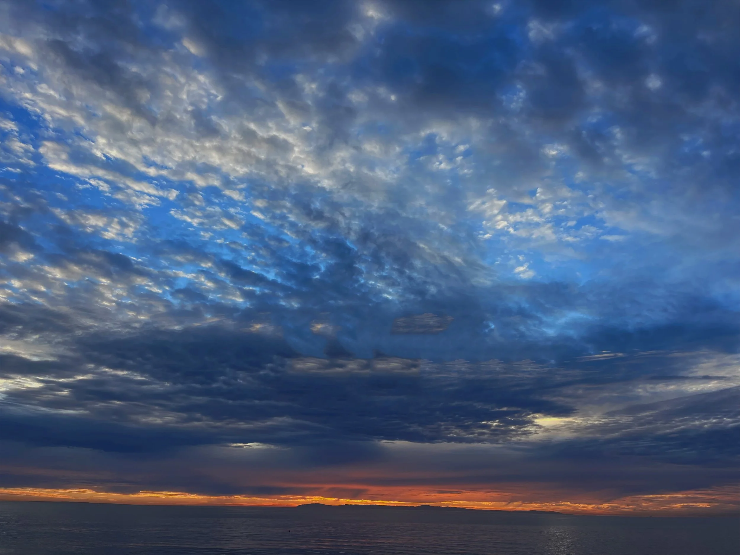 A scenic view of the sky at sunset with layered clouds, transitioning from dark gray at the top to vibrant orange near the horizon over the ocean.