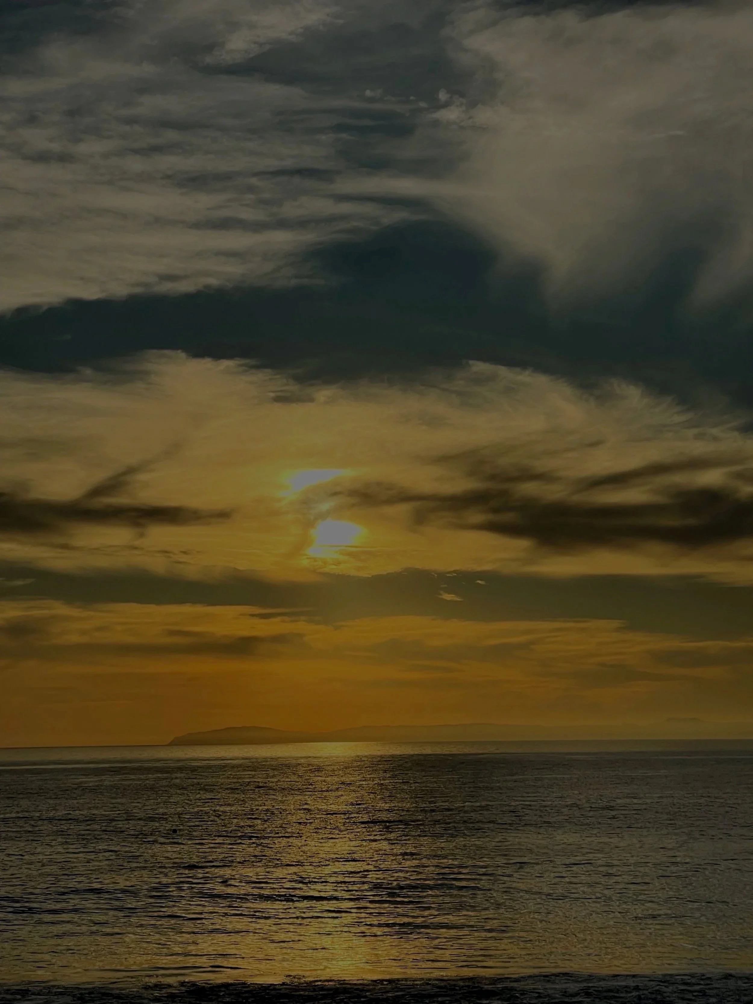 A sunset over the ocean with a partly cloudy sky, casting a golden glow on the water.