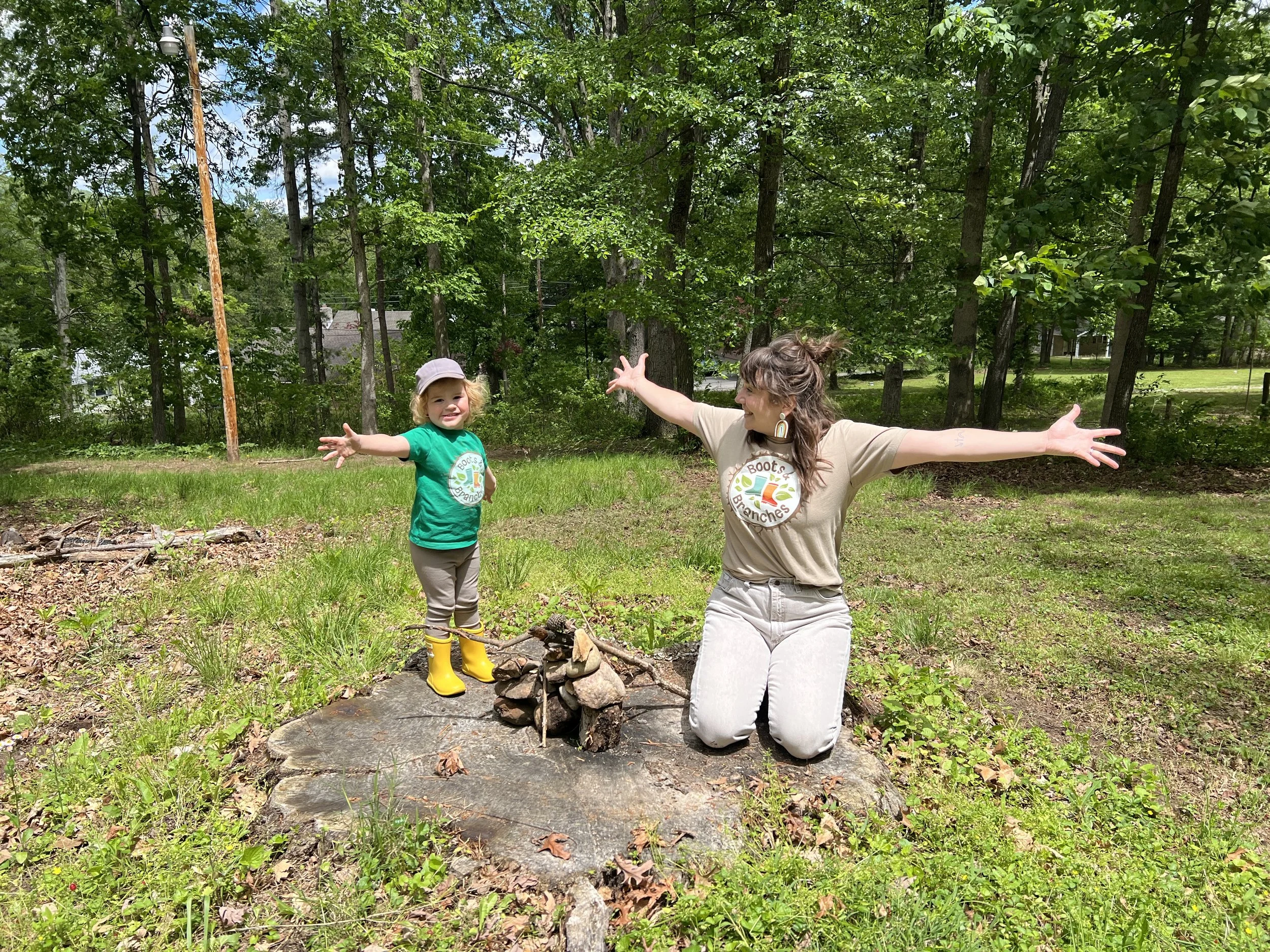 A woman and a young girl standing outdoors on a tree stump with their arms raised, surrounded by green grass and trees, sunny day.