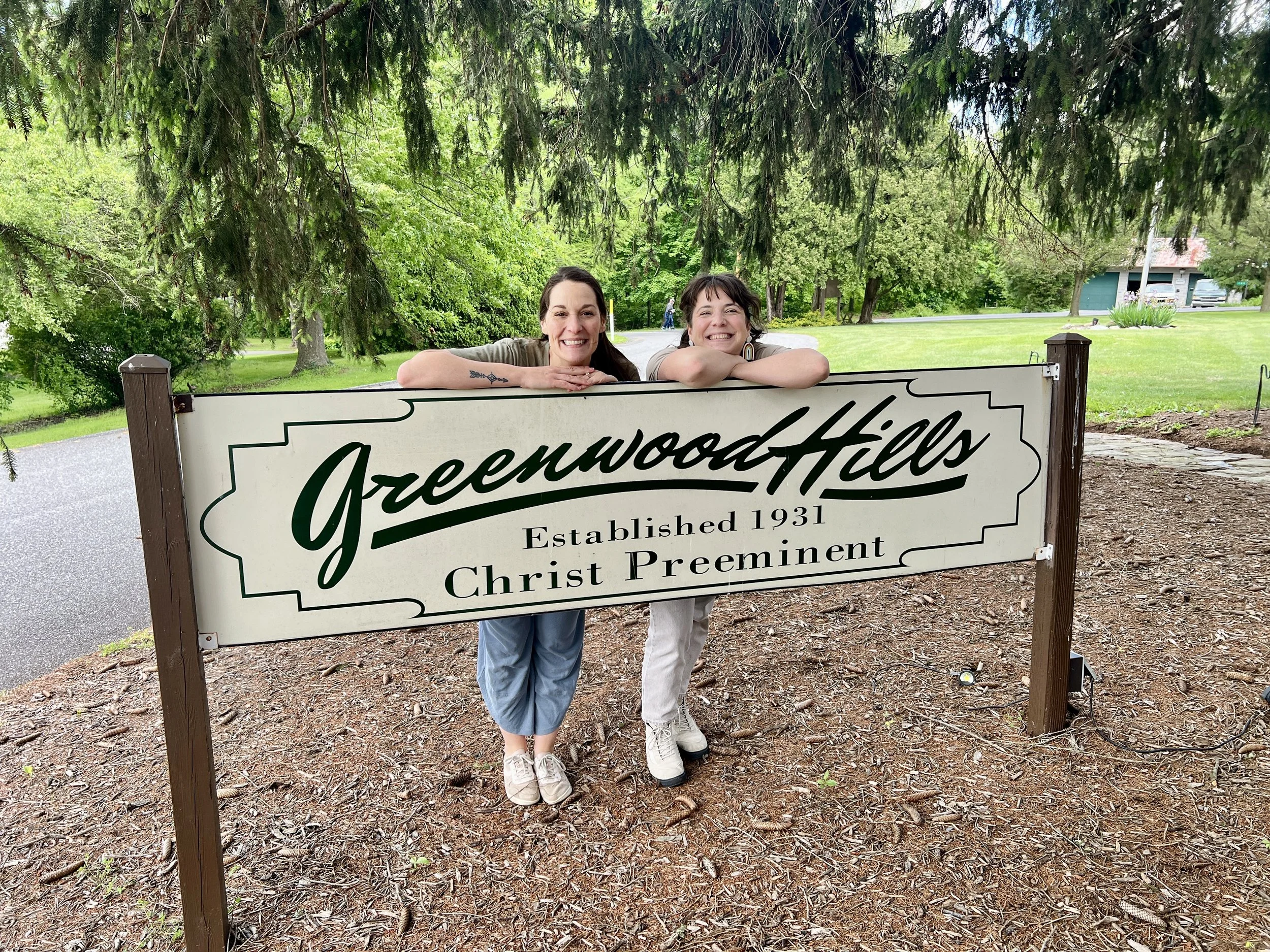 Two women leaning on a sign for Greenwood Hills with lush trees and greenery in the background.