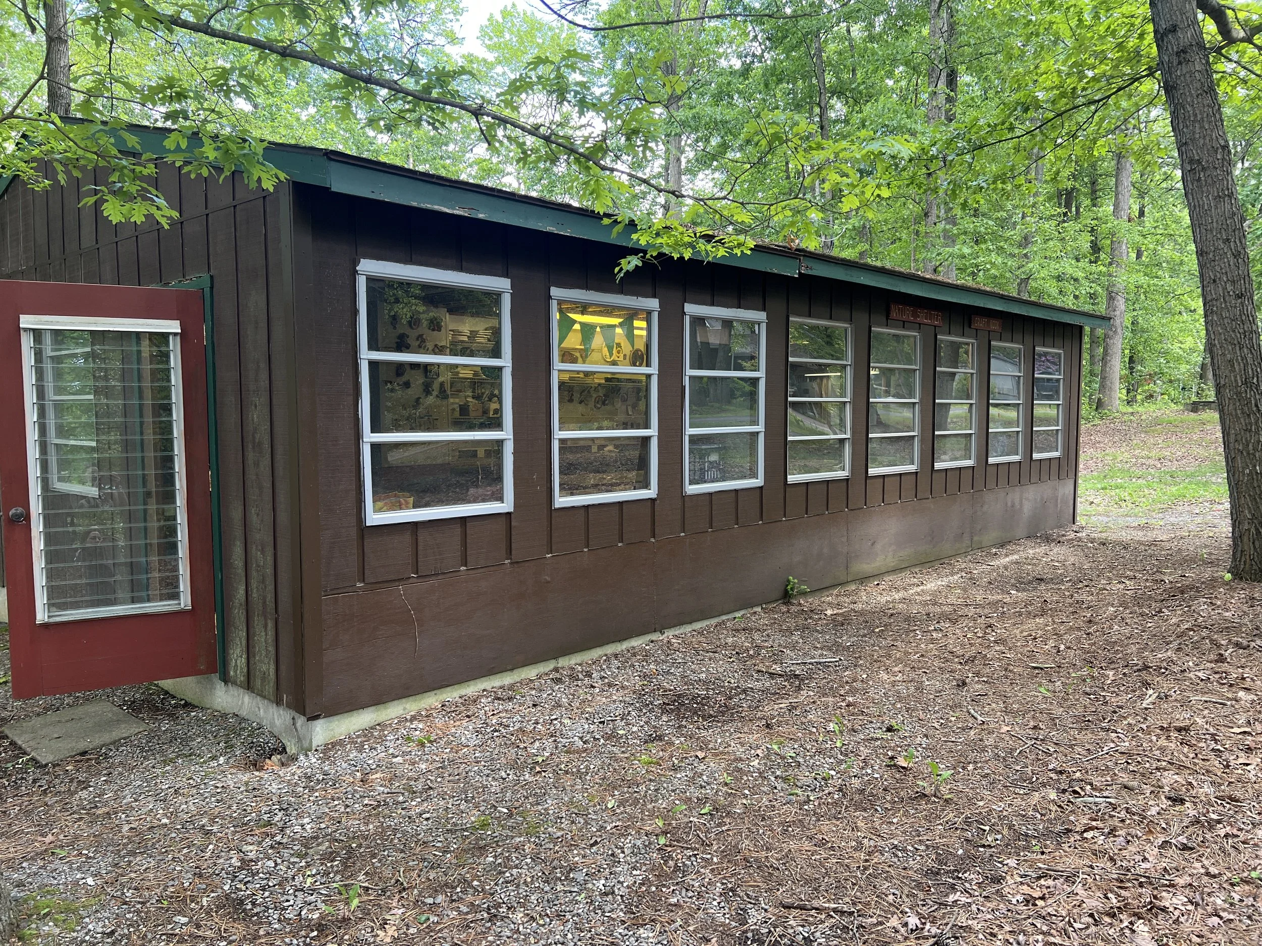 Brown wooden building with multiple windows and a red door located in a wooded outdoor setting with dirt and leaf-covered ground.
