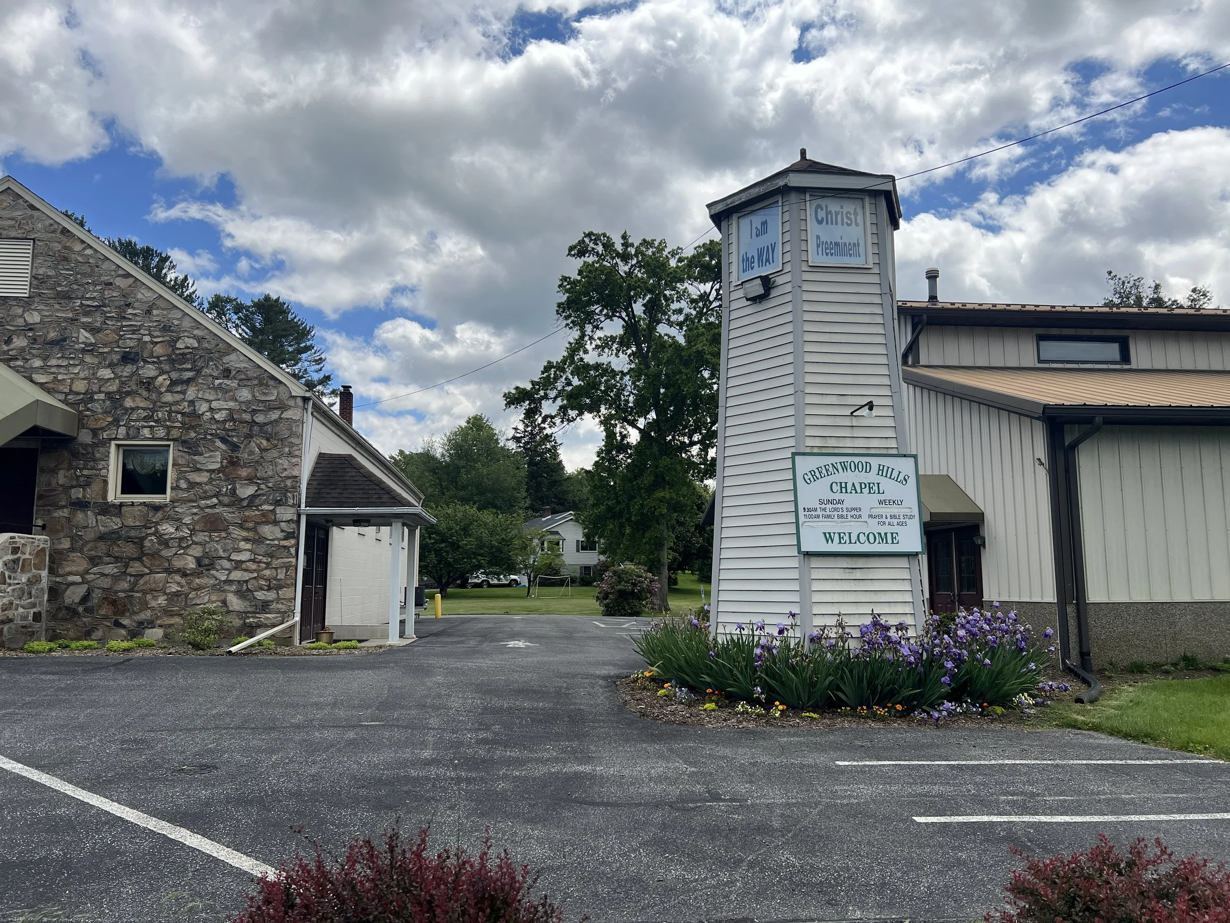 Church building with a white bell tower, a sign saying 'Greenwood Hills Chapel,' and a flower bed with purple flowers in front. The sign indicates worship service times and welcomes visitors.