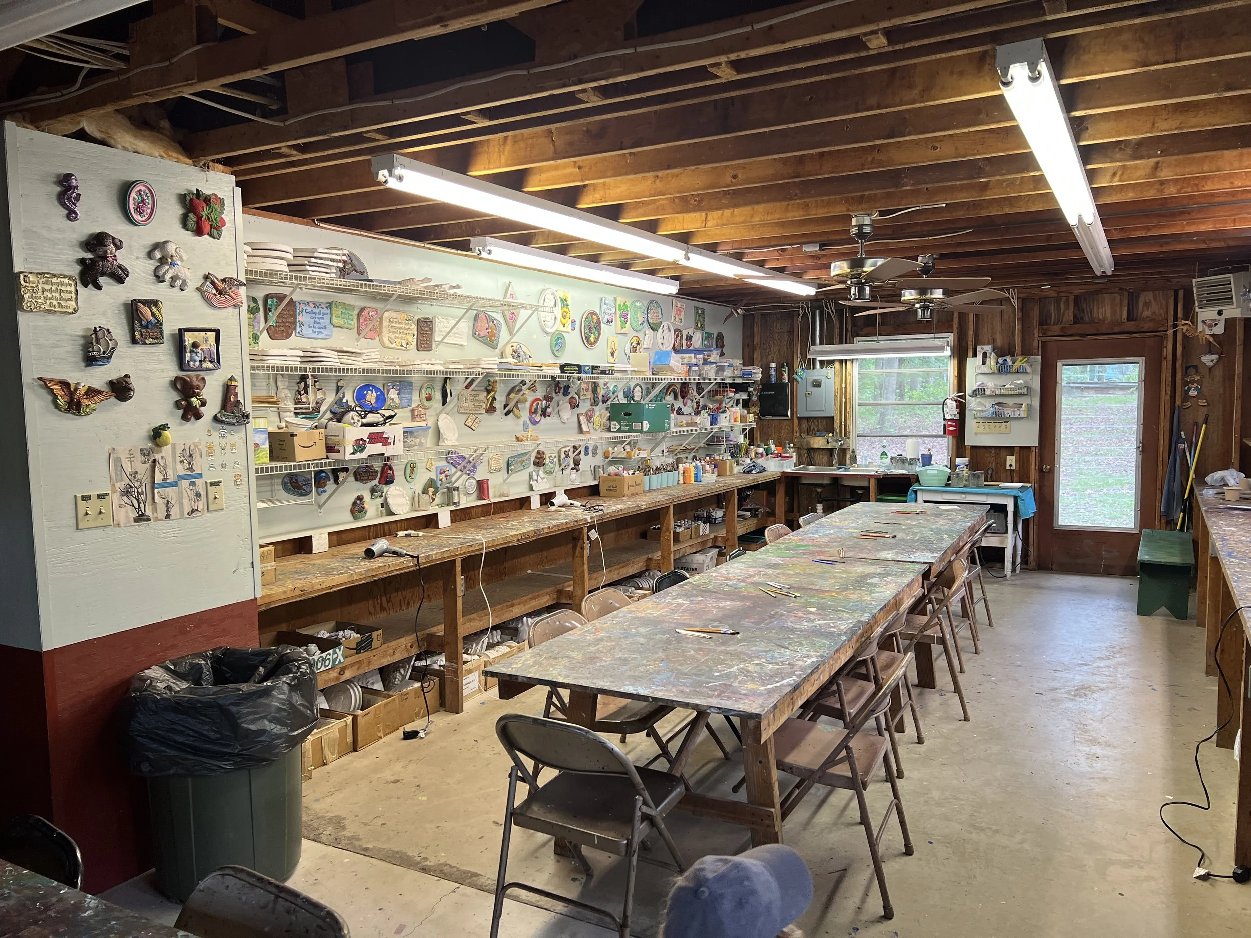 An art studio with a long painted worktable, shelves filled with various art supplies and decorations, and walls decorated with small colorful crafts and plaques. Large windows and a door provide natural light.