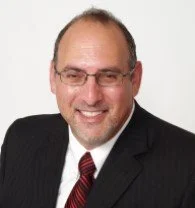 A smiling older man with glasses, a beard, and a mustache wearing a black t-shirt, a red cap, and large black headphones.