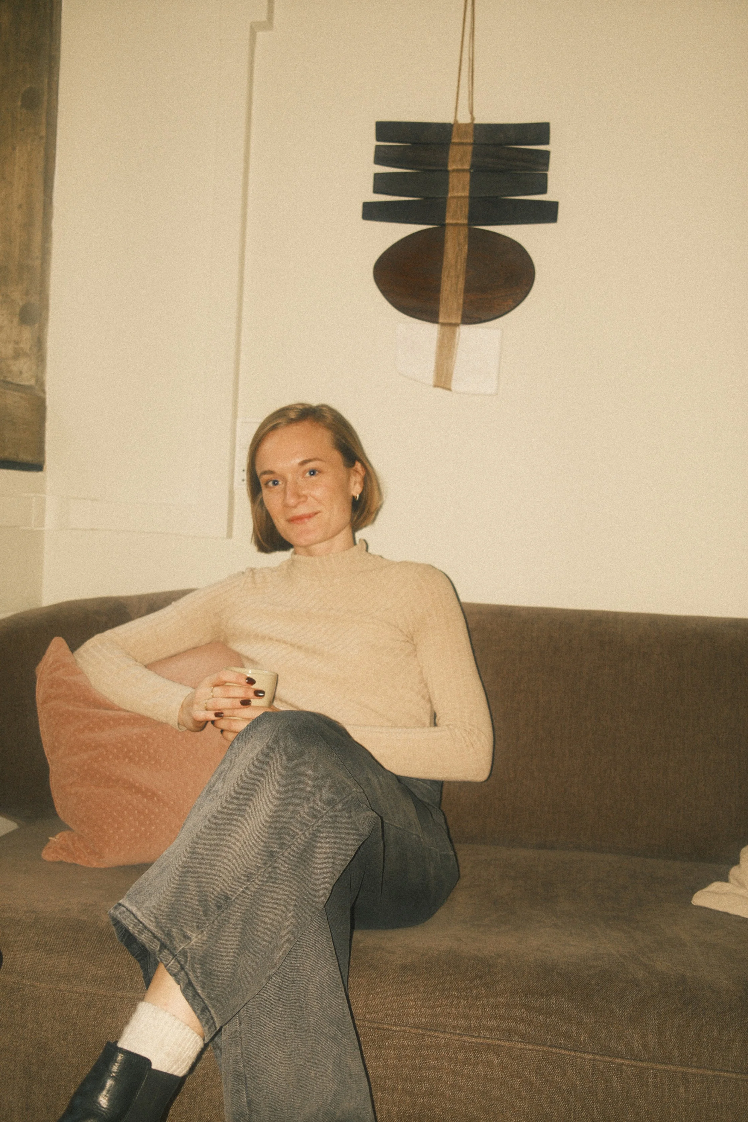 En kvinde sidder afslappet på en sofa, holder en kop. Hun har på en beige sweater, mørke bukser, beige sokker og sorte støvler. Der er en dekorativ vægkunst over hende.