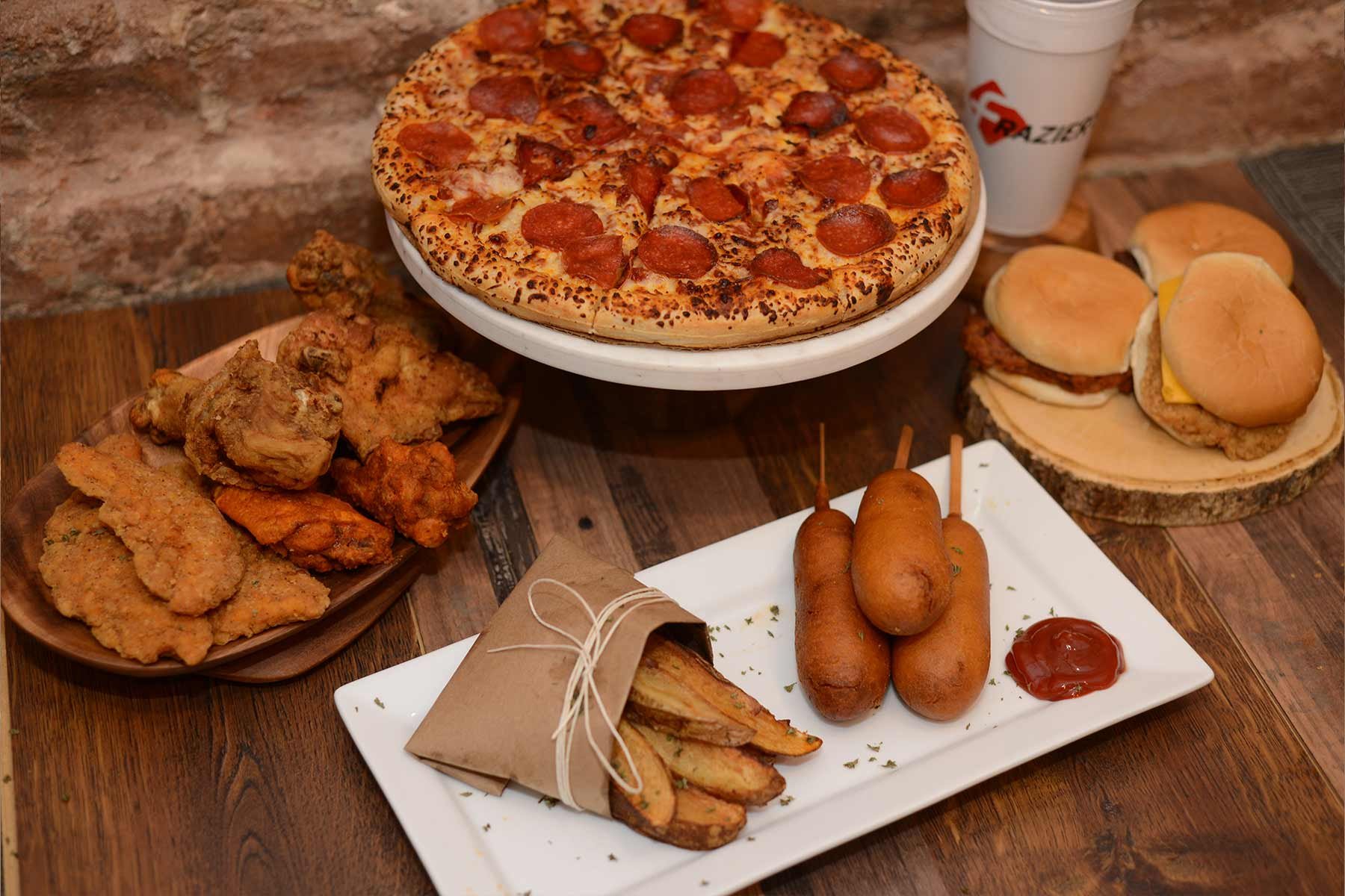 Assorted fast food including pepperoni pizza, fried chicken, burgers, corn dogs, and potato wedges on a wooden table.