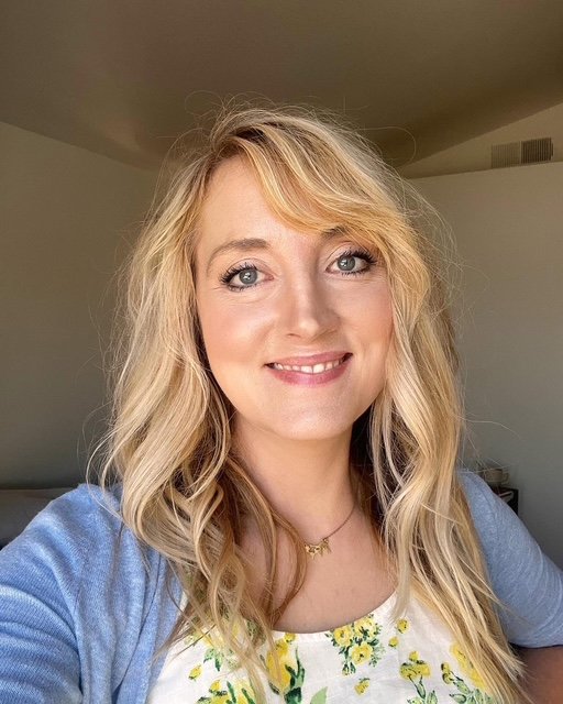 Close-up of a smiling woman with long blonde hair, blue eyes, and wearing a dark blue top, standing against a plain light background.