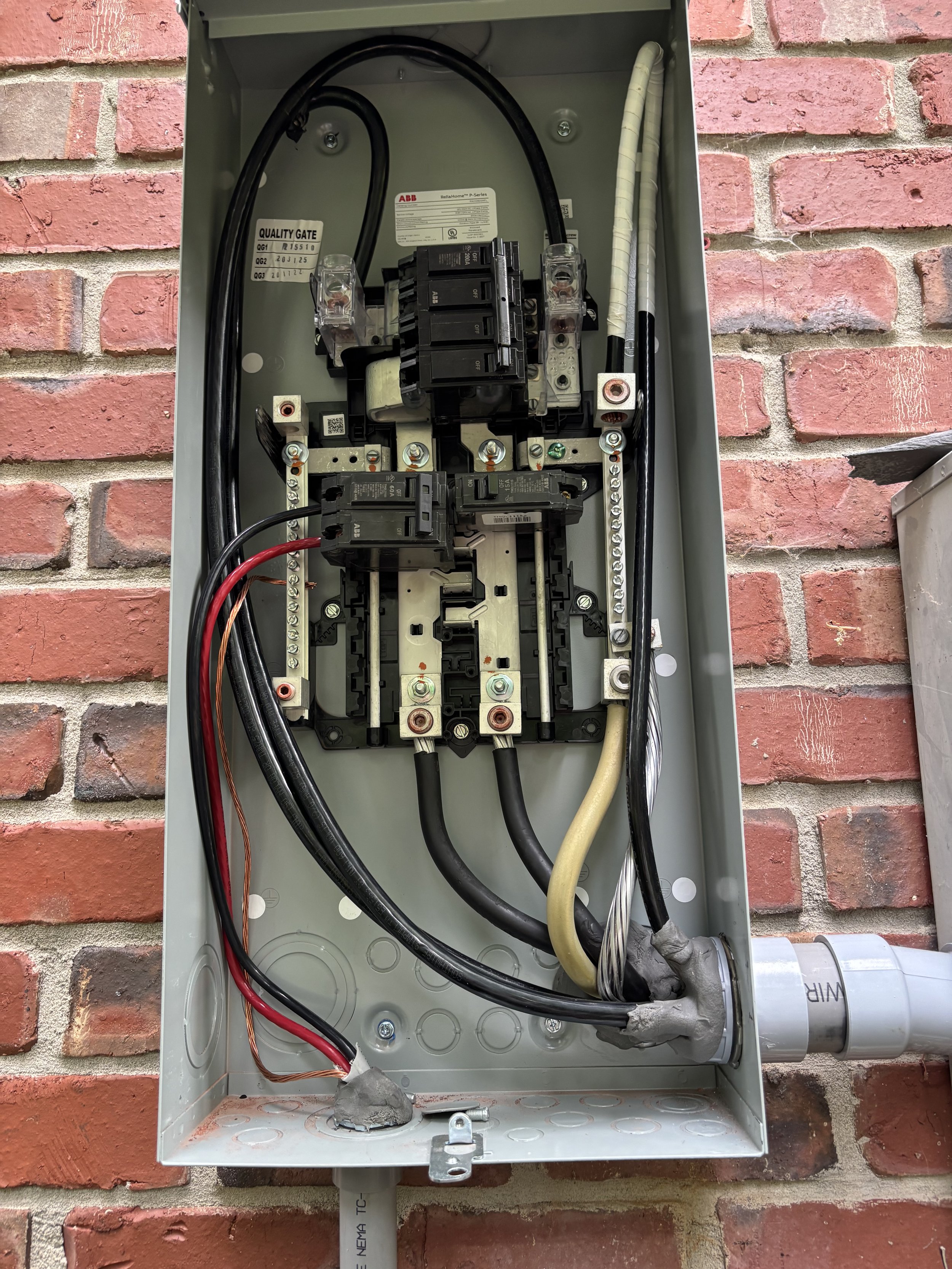 Open electrical control panel with various wires, relays, and circuit breakers mounted inside, attached to a brick wall.