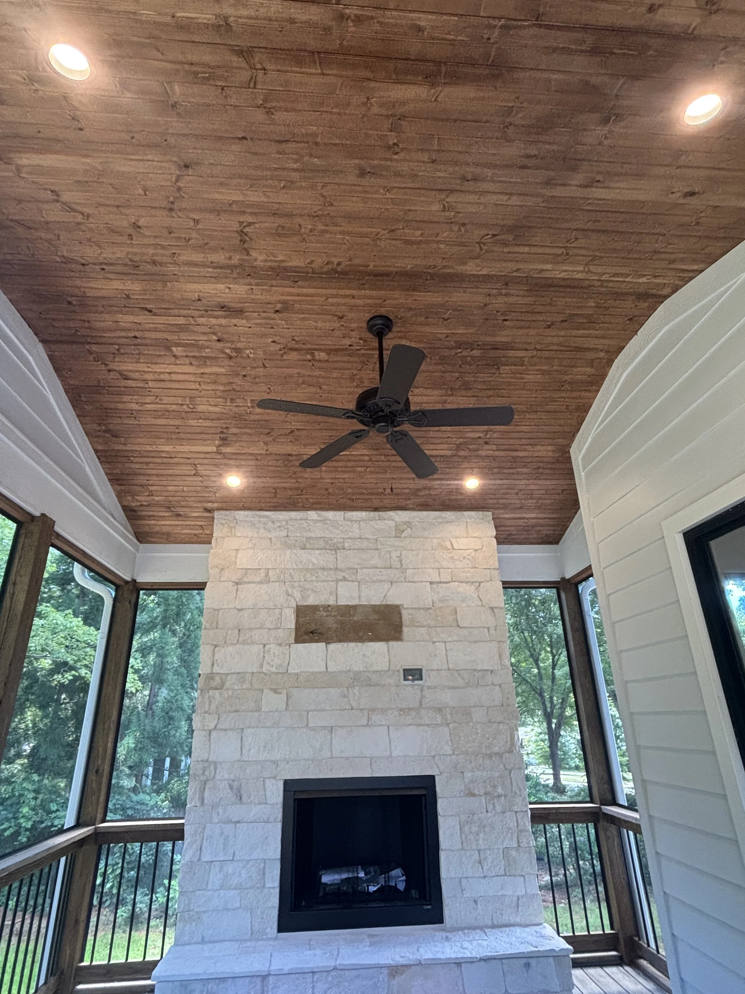 A ceiling with wooden planks, four recessed lights, and a ceiling fan. Below is a stone fireplace with a wooden patch and an electrical outlet. Large windows on both sides offer a view of green trees outside.