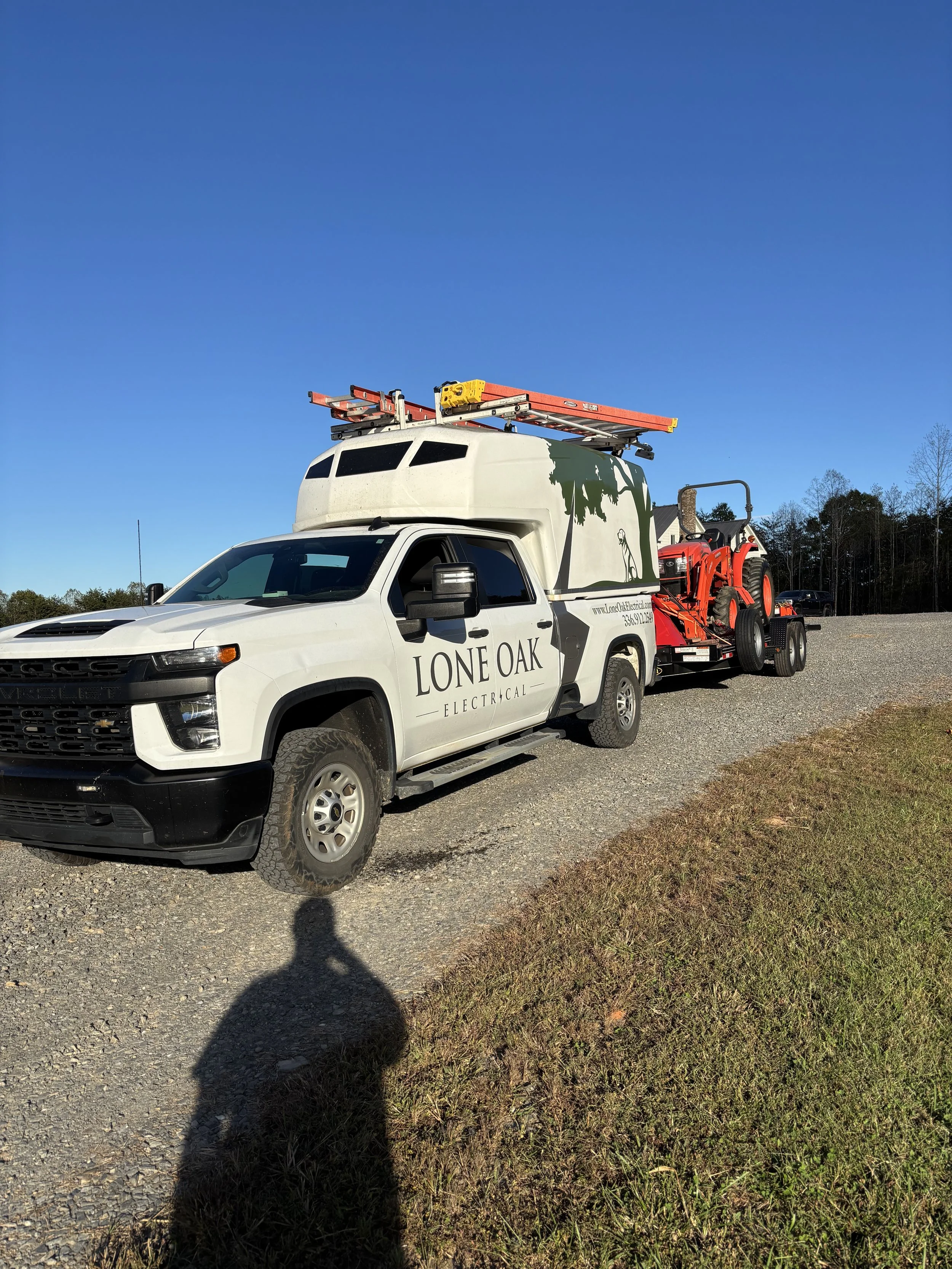 White utility truck with 'Lone Oak Electrical' written on the side, carrying ladders and a red tractor loader, parked on gravel with a grassy area and trees in the background.