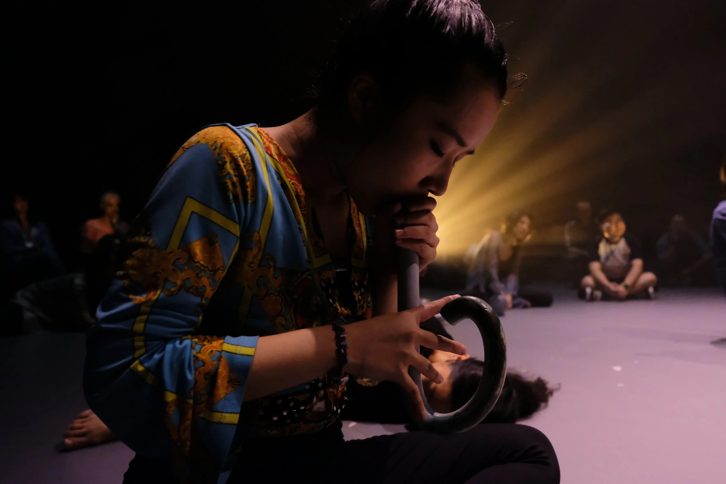 Sing-Yatwith dark hair and colorful clothing kneels down while holding a ceramic flu, with her head bowed and eyes closed. She is in a dimly lit room with a group of people sitting in the background, suggesting a relaxed or meditative group activity.