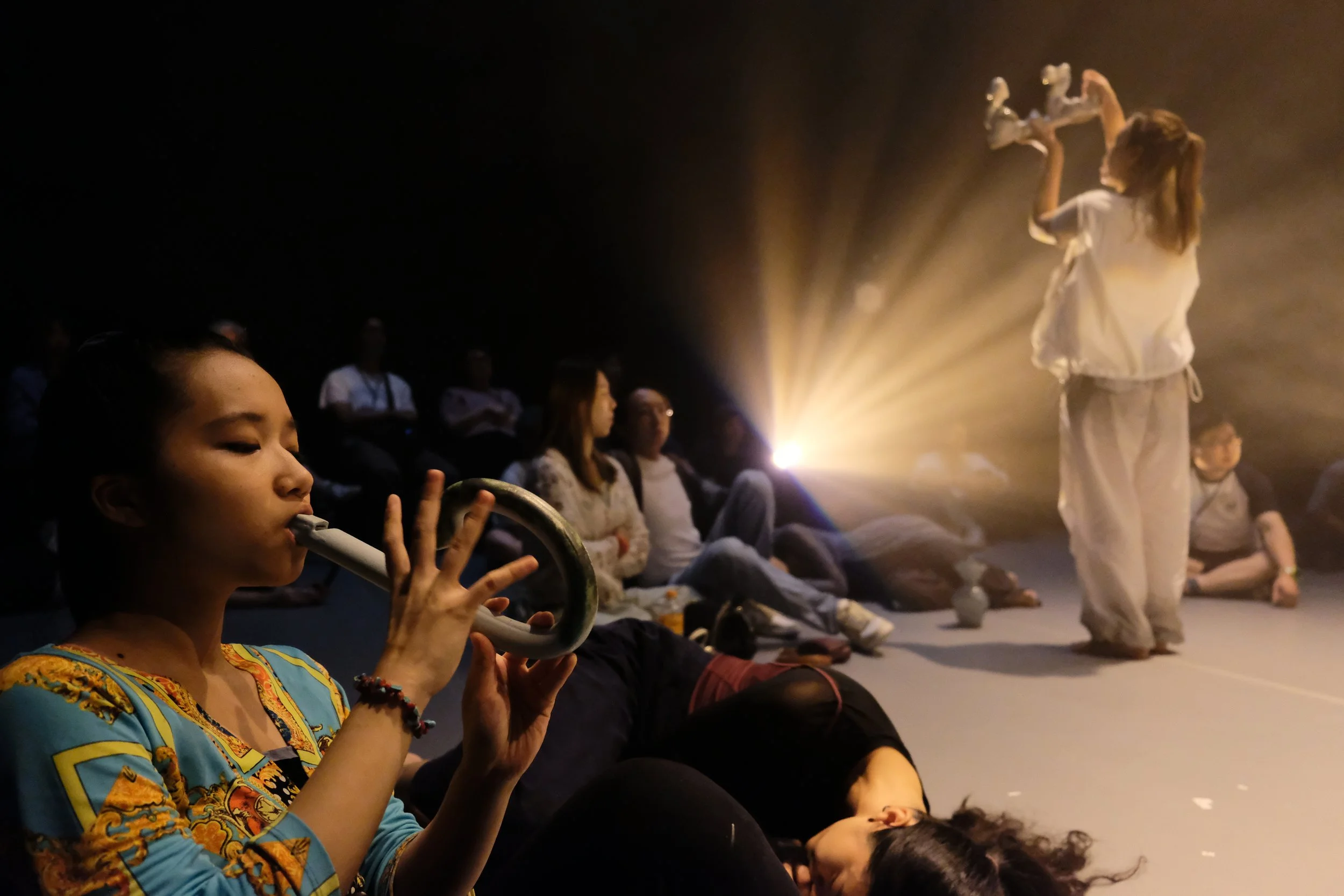 Sing-Yat in a colorful dress plays a ceramic flute while sitting on the floor, surrounded by an audience of people, with a person standing in the background illuminated by a warm light.