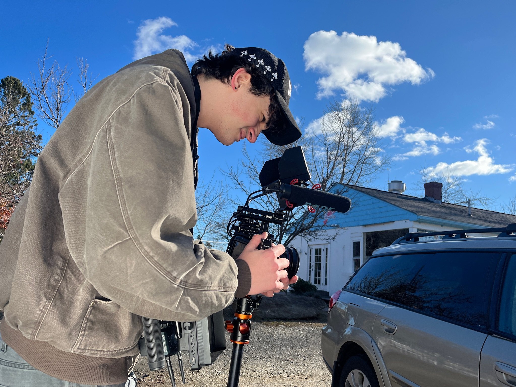 Young man operating a professional video camera outdoors in front of a house, with a clear blue sky and some trees in the background.