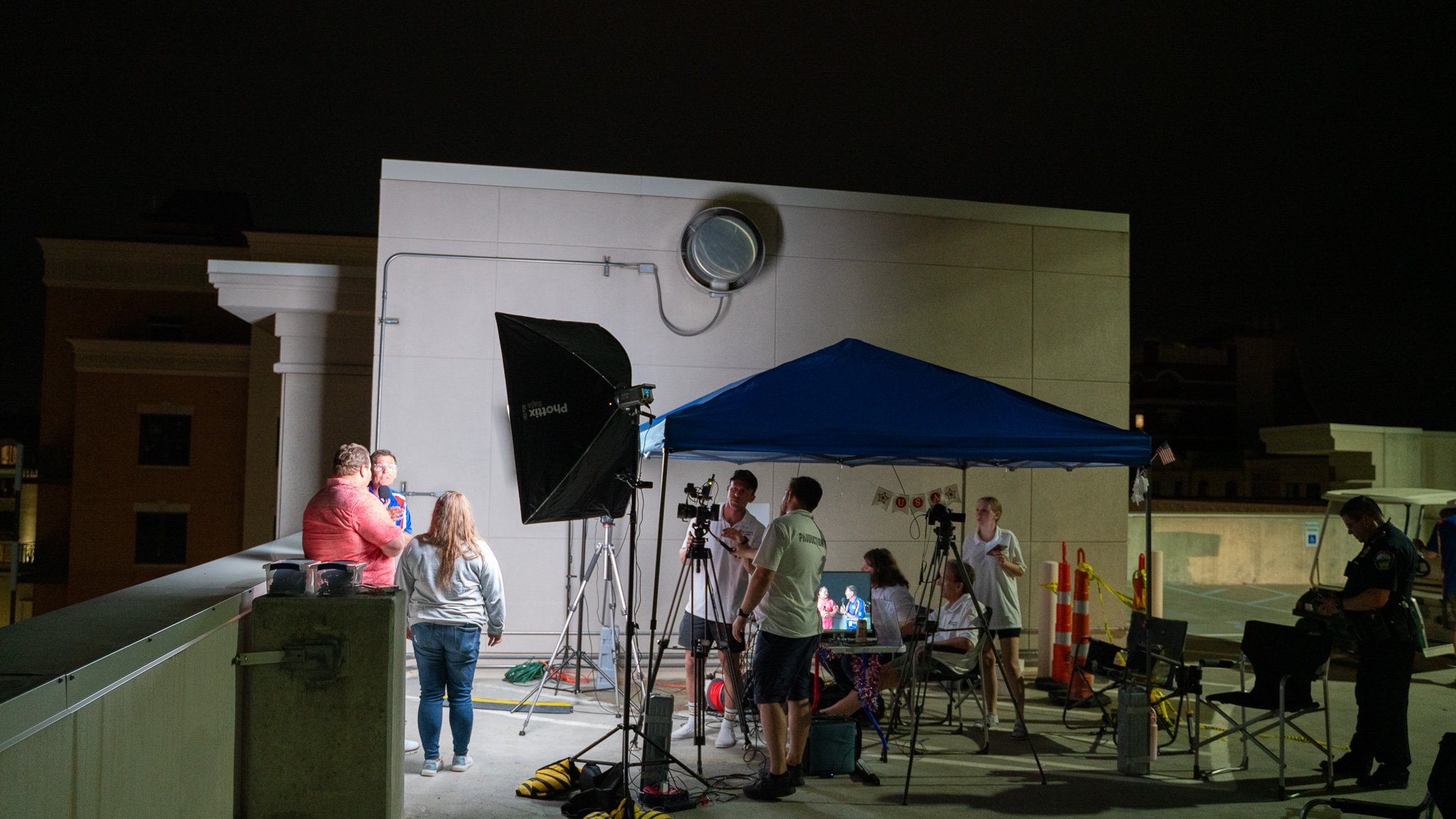 Nighttime outdoor scene with a movie or TV show being filmed under a blue canopy, with cameras, lighting, crew members, and actors on set.