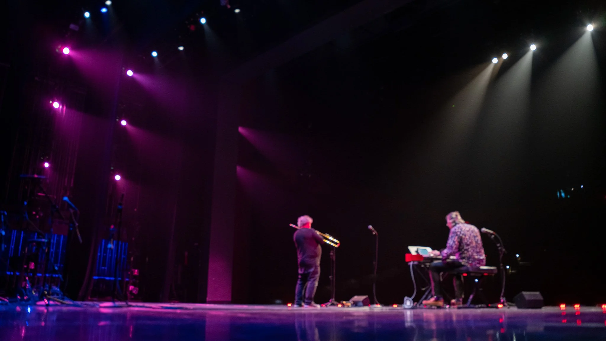 Musicians performing on stage with purple and white lights, one playing keyboard and another holding a trombone.