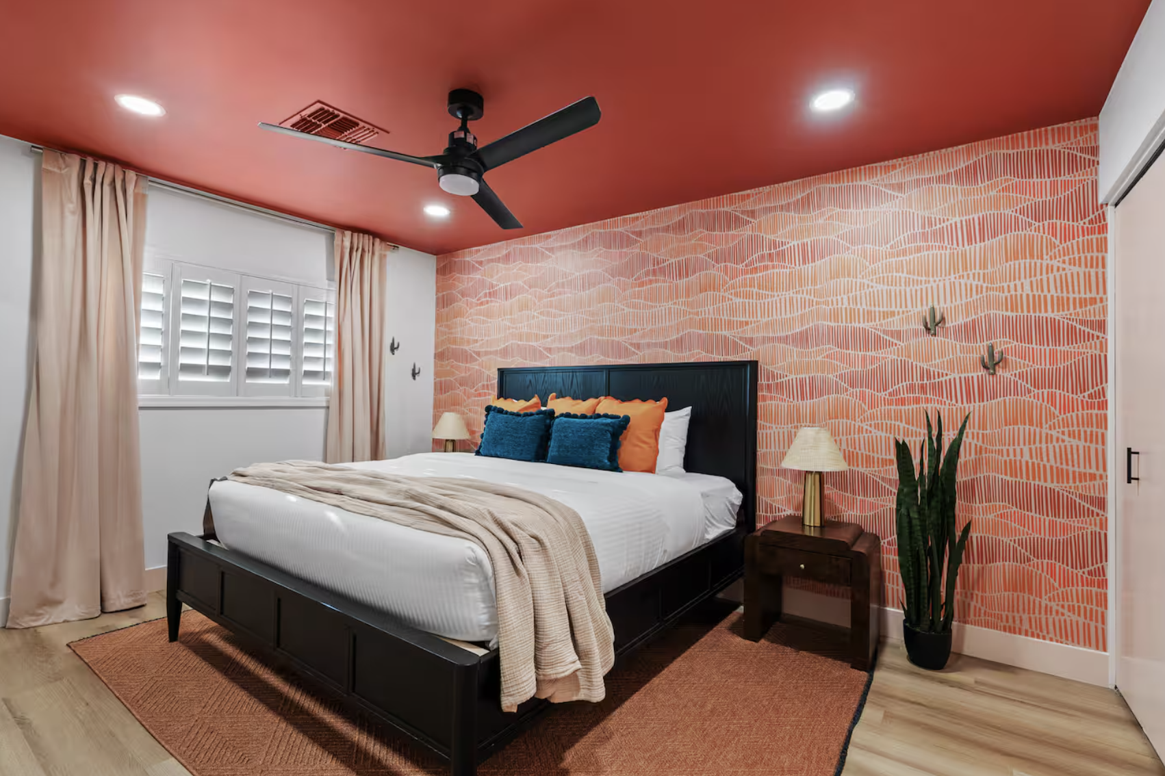 Bedroom with a bed, orange and blue pillows, beige blanket, wooden nightstand, plants, lamps, and a window with shutters, featuring a coral and pink patterned accent wall and ceiling.