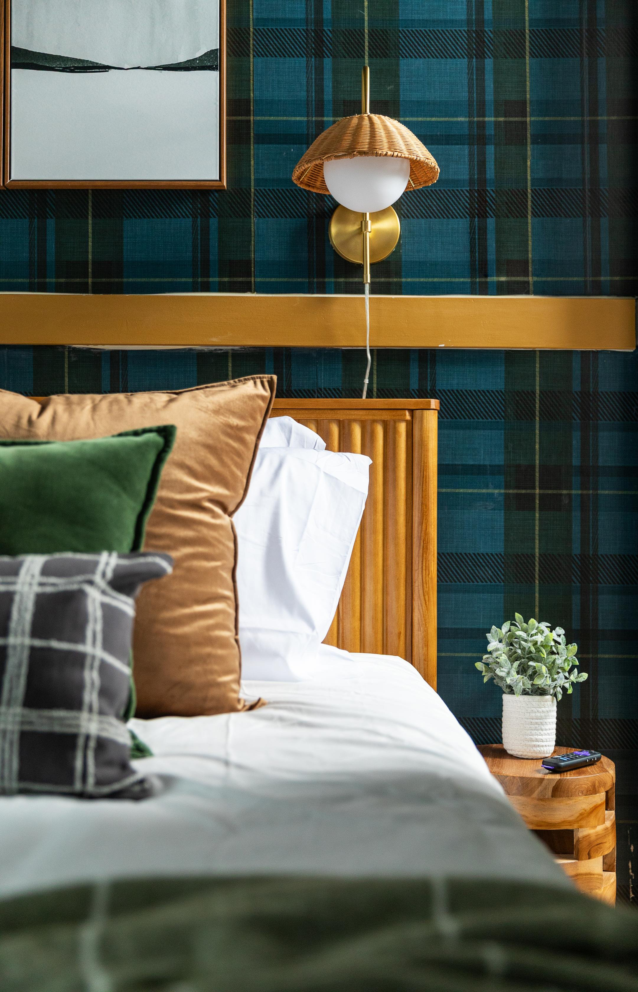 Close-up of a neatly made bed with assorted pillows in brown, green, and plaid patterns, set against a dark green plaid wallpaper. On the bedside table, there's a potted plant, a remote control, and a round wooden table.