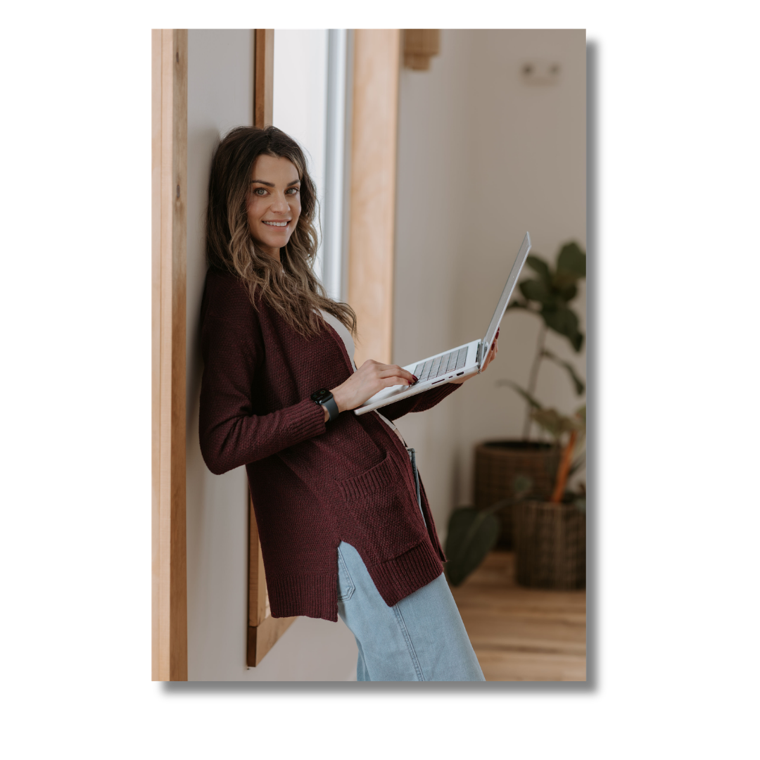 A young woman with long wavy hair smiling while standing against a wall and holding a laptop.