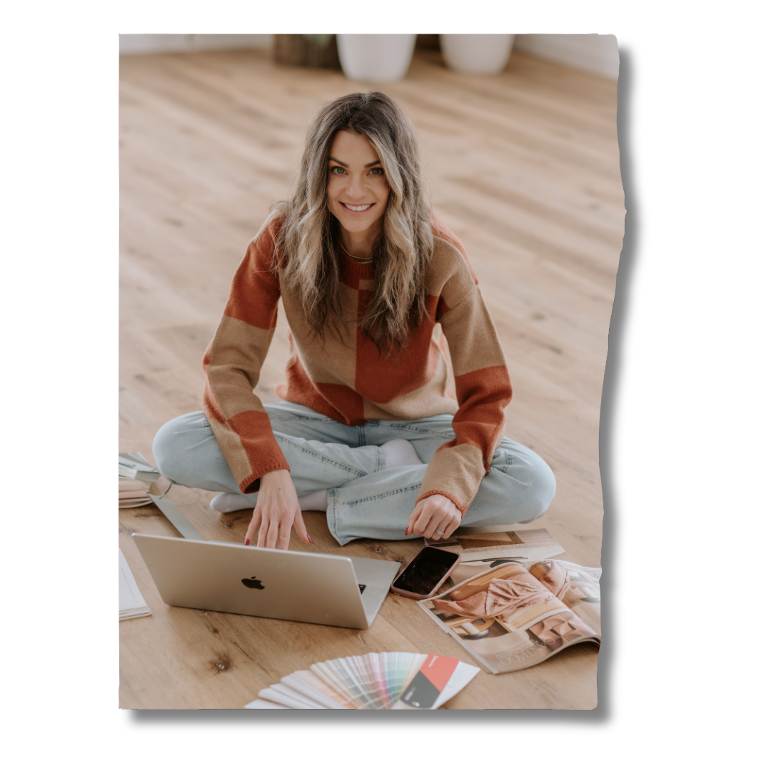 Young woman sitting cross-legged on wooden floor with a laptop, smartphone, open magazines, and painted color swatches, smiling at the camera.