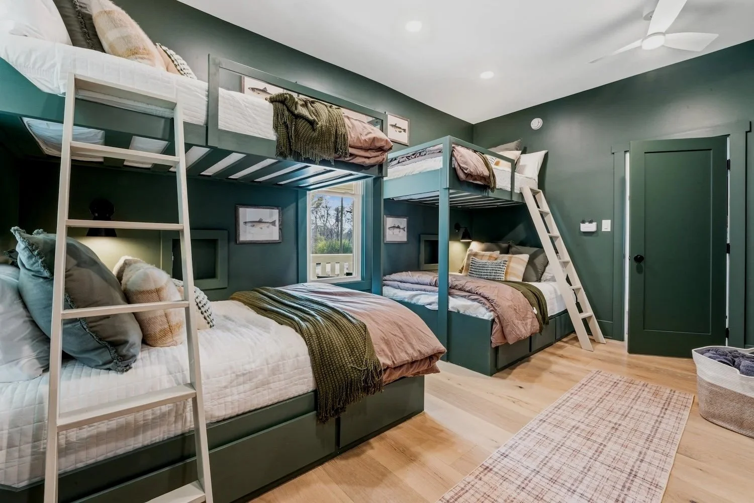 Room with four beds arranged as two sets of bunk beds with ladders, green walls, framed aquatic animal pictures, window showing trees outside, wooden floor, ceiling fan, and a patterned rug.