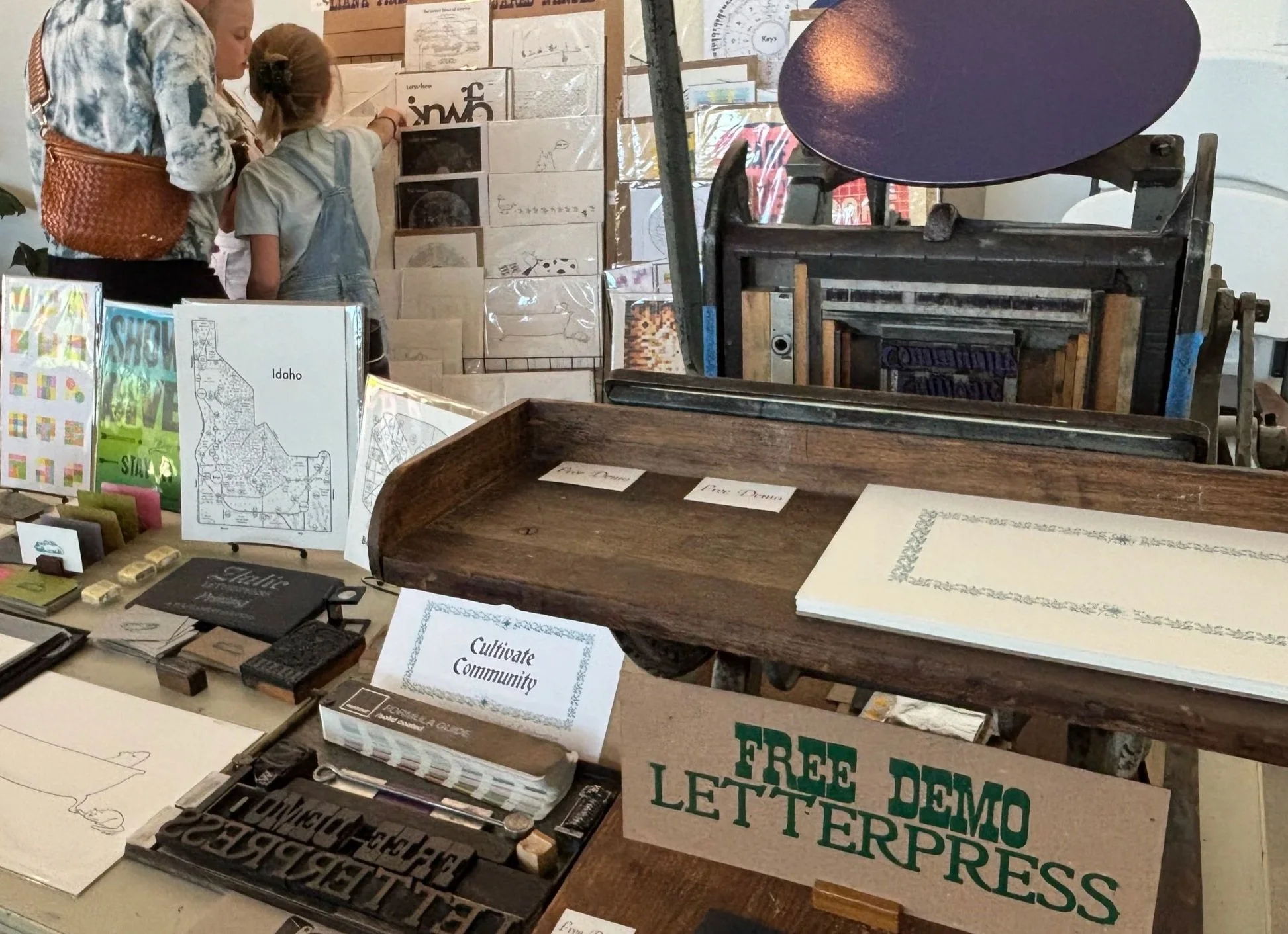 A display of various print materials including books, maps, and signs promoting community and democracy, with two children and an adult engaging with the items. The scene appears to be in a community event or educational setting.