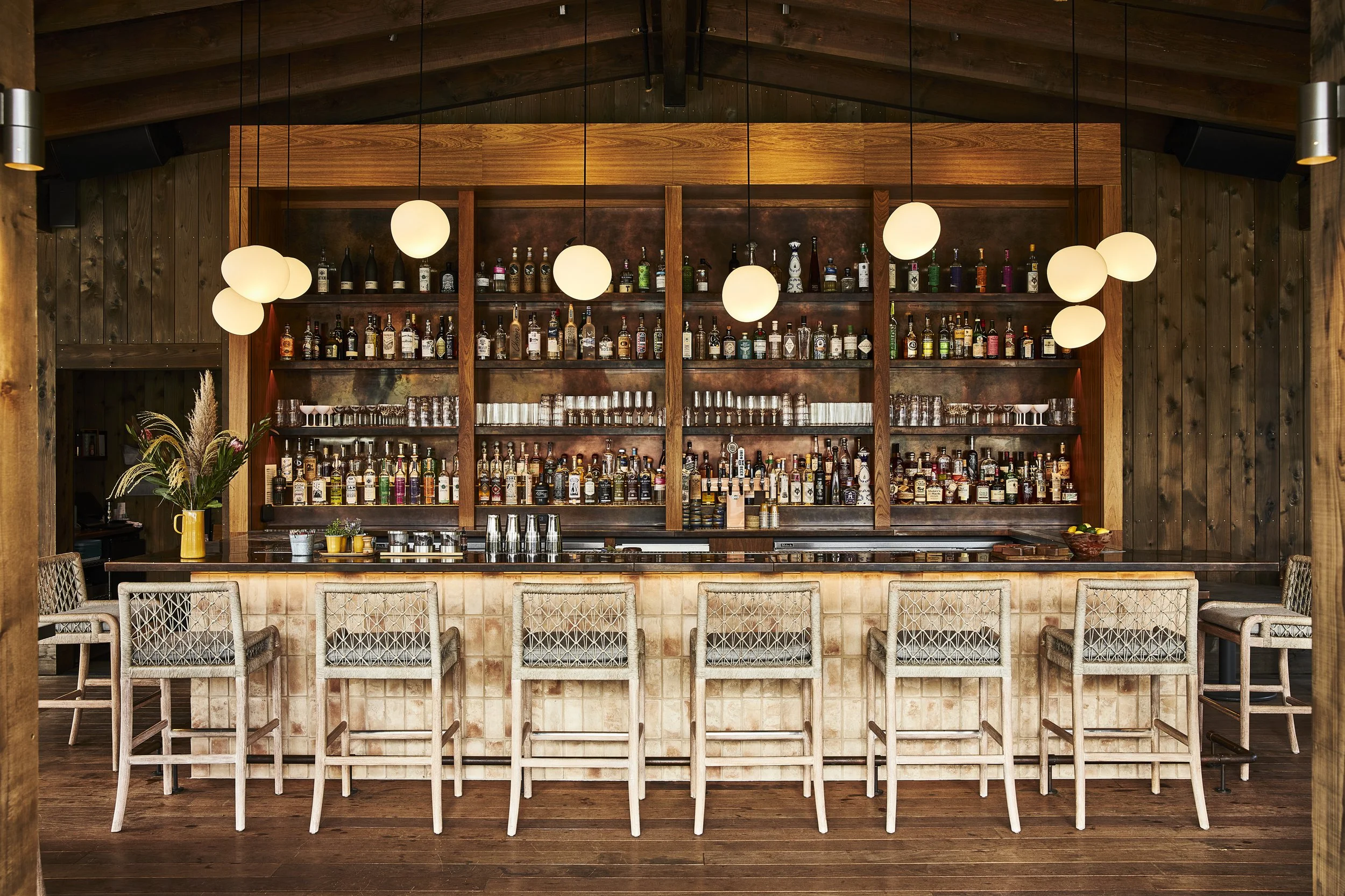 A bar area with wooden decor, glass bottle shelves, hanging globe lights, and eight bar stools in front.