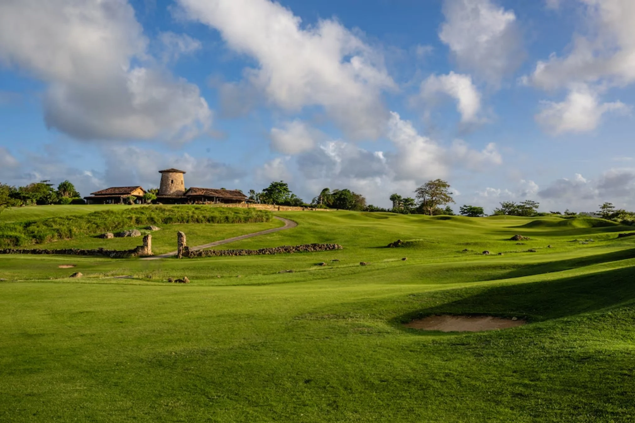 A lush green golf course under a partly cloudy sky with a historic stone building in the background.