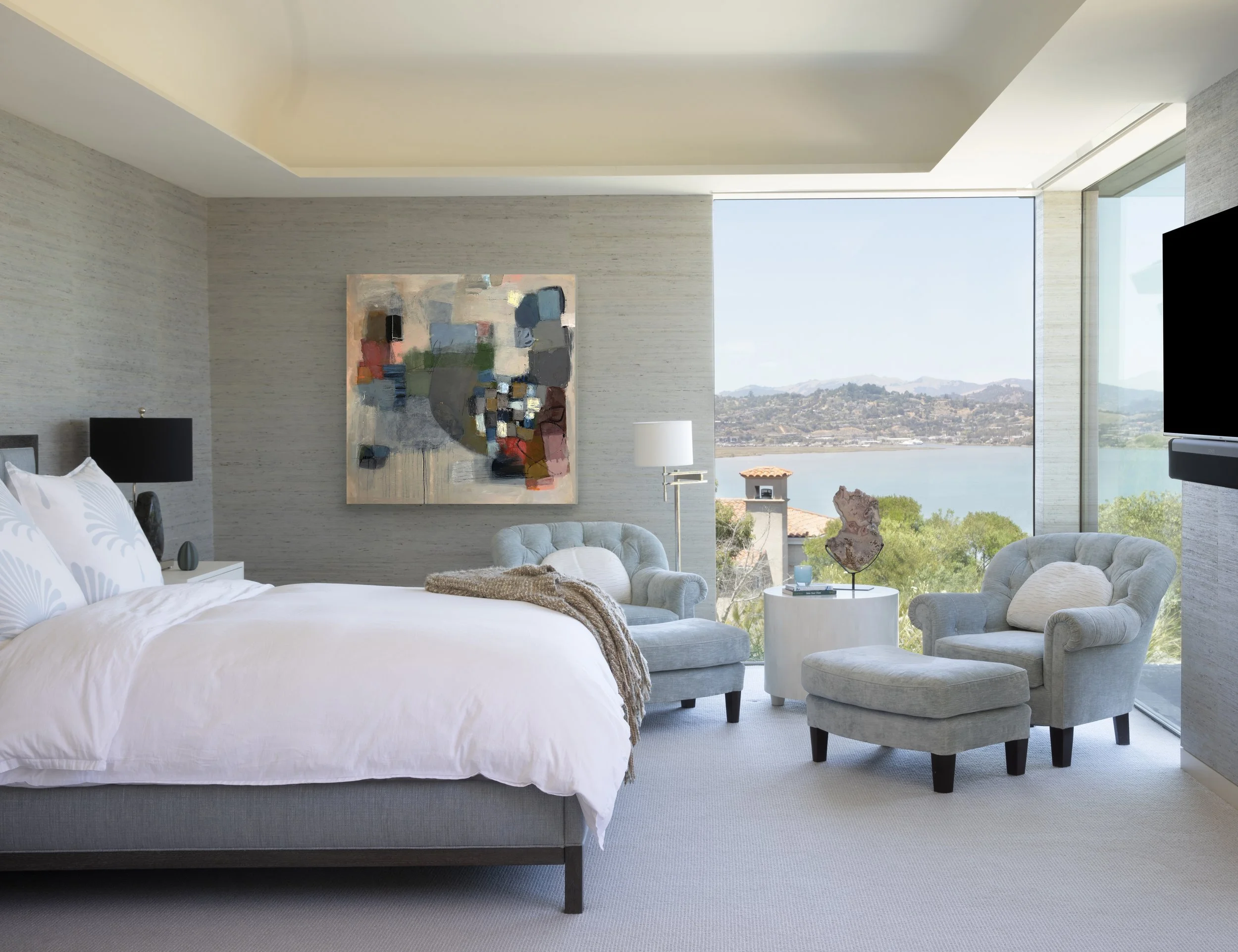 Modern bedroom with a large bed, abstract artwork on the wall, two armchairs, a chaise lounge, a small white side table with a sculpture, and a large window showing a scenic view of a body of water and hills.