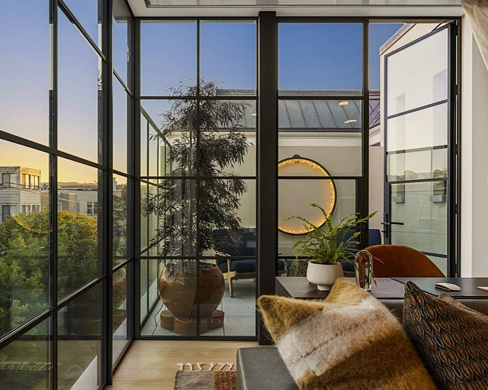 View through glass walls of a modern room with indoor plants, a round mirror with warm lighting, and an outdoor balcony with trees and buildings in the distance.