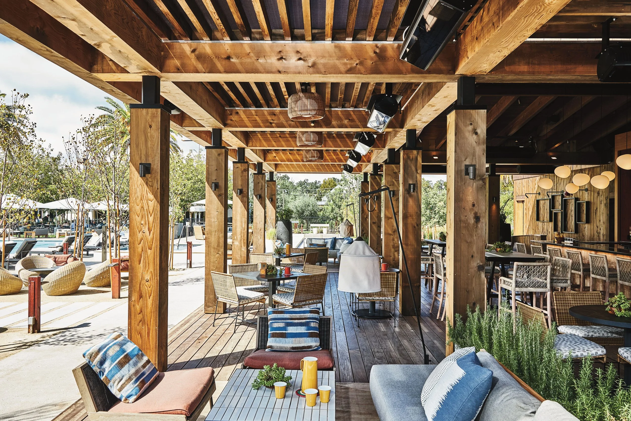 Outdoor patio with wooden beams and furniture, including chairs, sofas, and tables, under a wooden ceiling with hanging lights, surrounded by trees and pool area in the background.