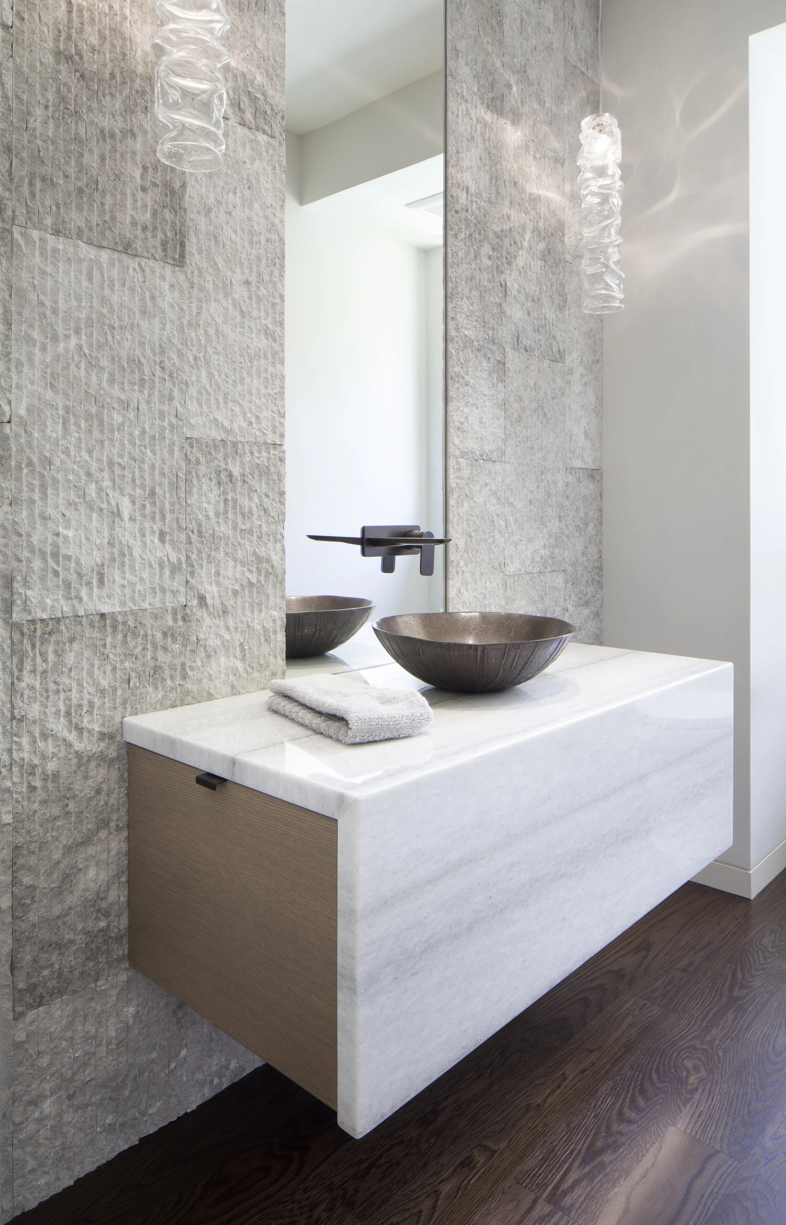 Modern bathroom vanity with two dark metal bowls, a gray towel, a white marble countertop, and textured stone wall, illuminated by pendant lights.
