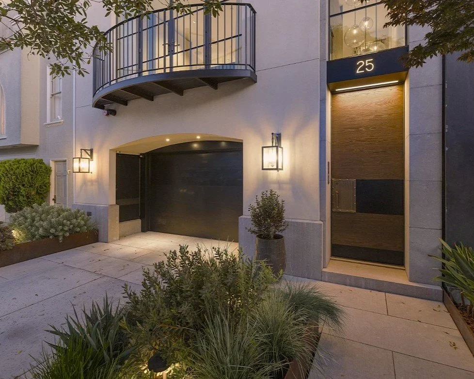 Modern residential building entrance with black garage door, wooden front door, house number 25, outdoor lighting, and landscaped plants.