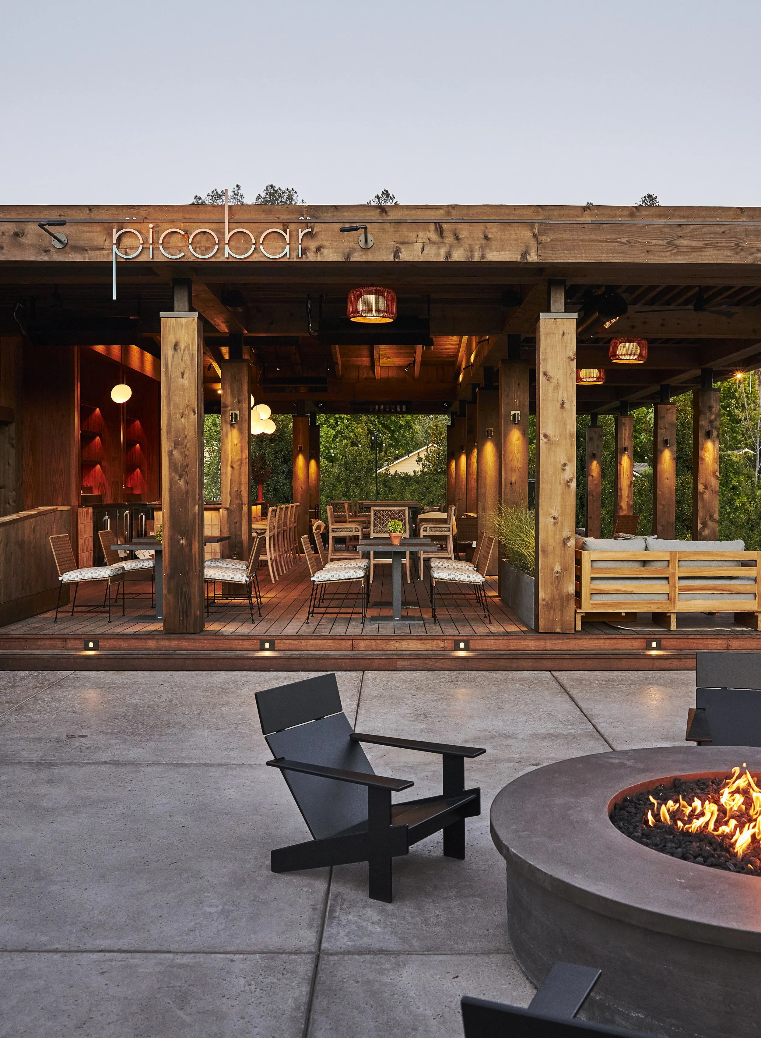 Exterior view of a modern restaurant named 'picobar' with a cozy outdoor seating area featuring fire pit, chairs, and tables under a wooden structure with warm lighting.