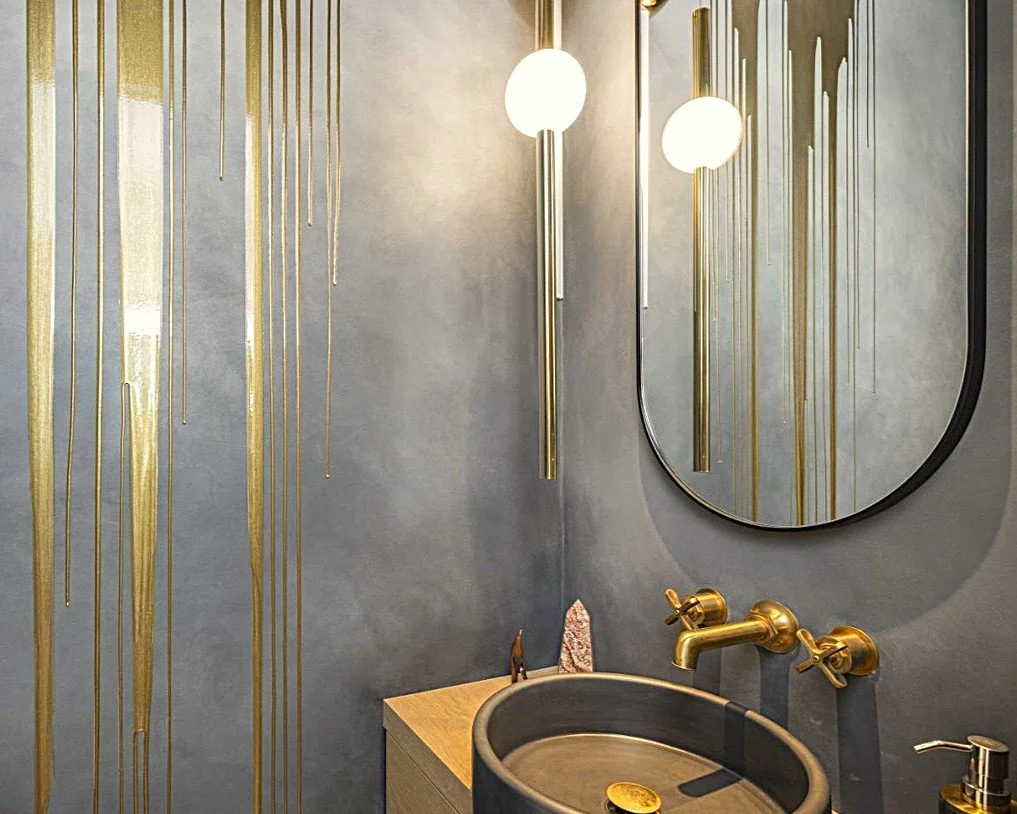 Modern bathroom with grey walls, gold accents, round mirror, and unique sink with gold faucet.