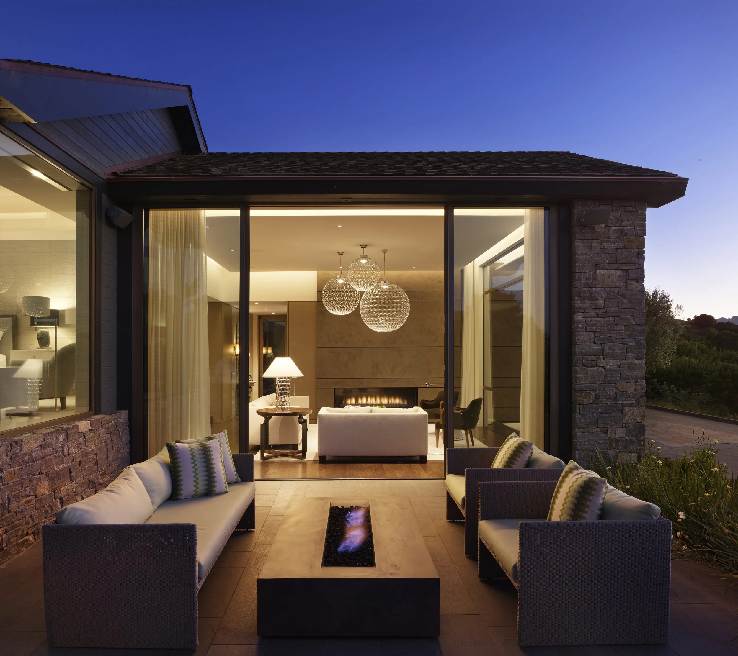 Outdoor patio with sofas and a fire pit, view into a modern living room with a fireplace, chandelier, and large windows, during twilight.