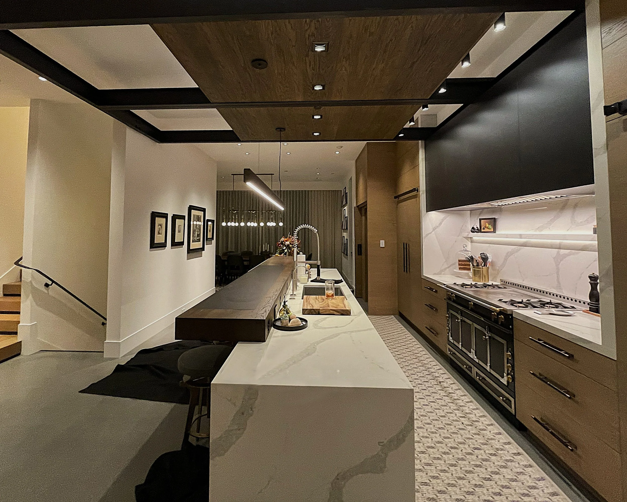 Modern kitchen with a marble island, wooden accents, and a gas stove with an oven underneath. The ceiling has a wooden panel with built-in lighting, and the walls are decorated with framed artwork.