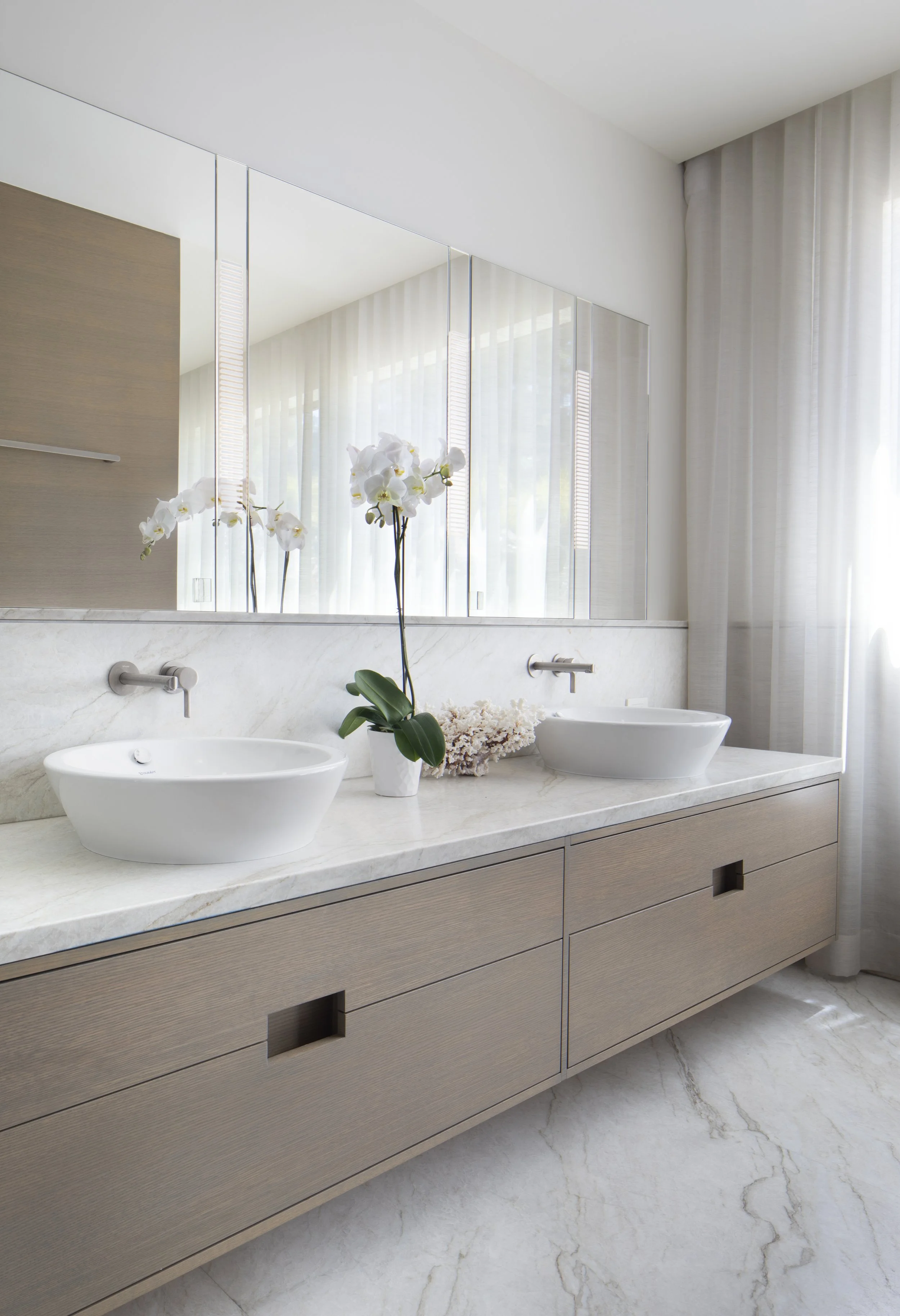 Modern bathroom with double vessel sinks, a large mirror, beige cabinetry, orchid plant, and natural light with sheer curtains.