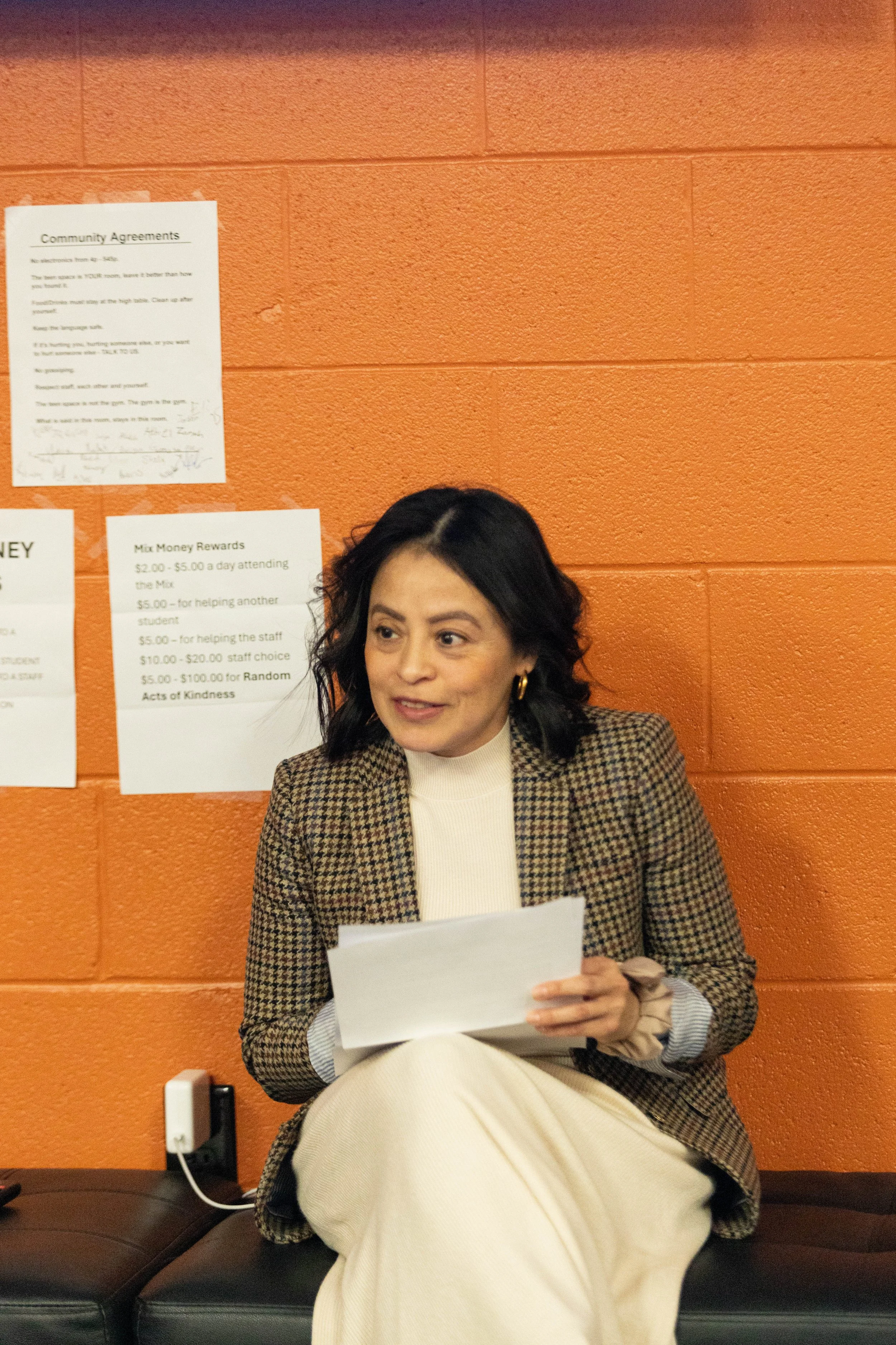 A woman with dark wavy hair wearing a checked blazer and cream-colored skirt, sitting on a black bench against an orange wall, holding a piece of paper.