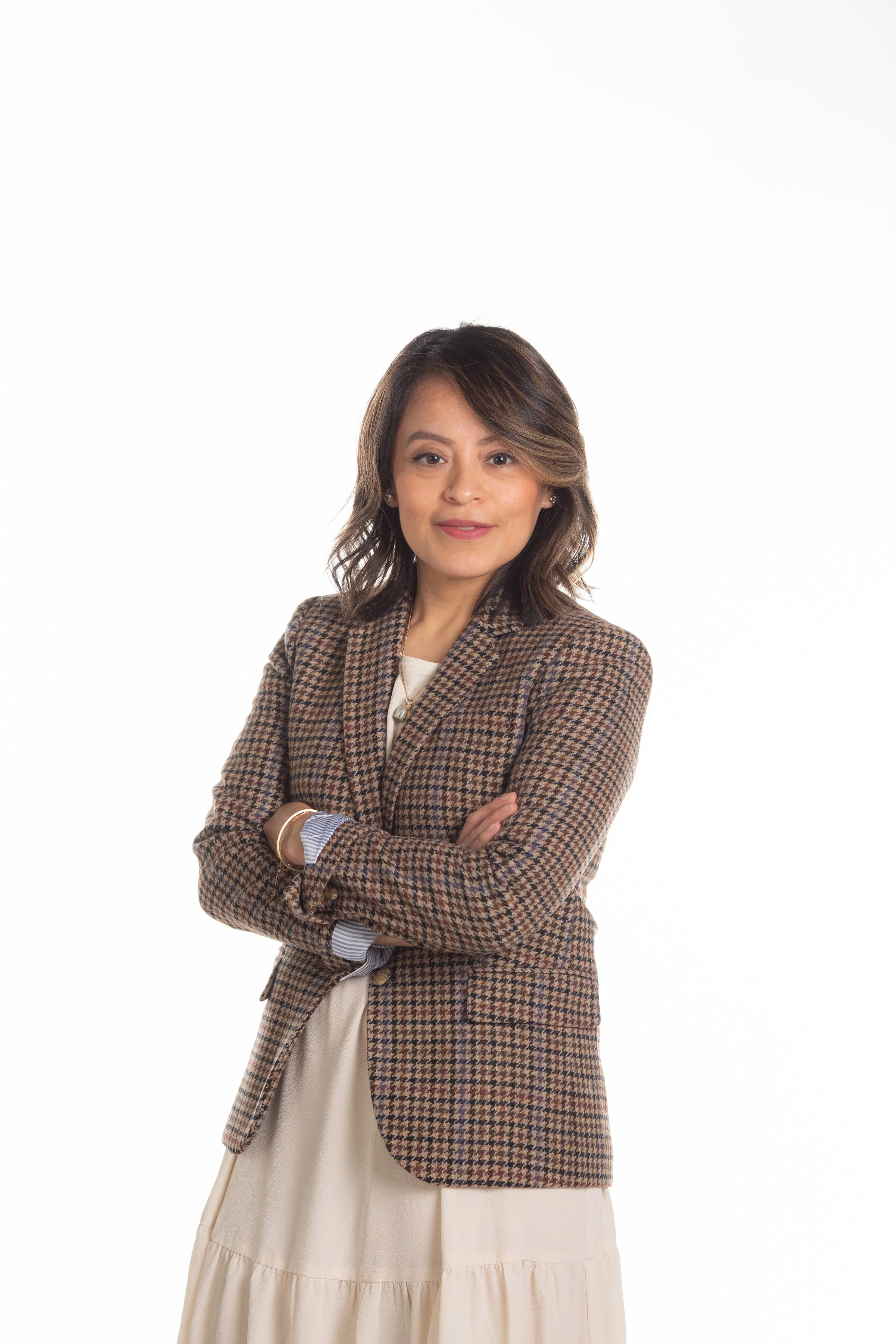 A woman with shoulder-length wavy hair wearing a brown checked blazer, white top, and beige skirt, standing with arms crossed against a plain white background.