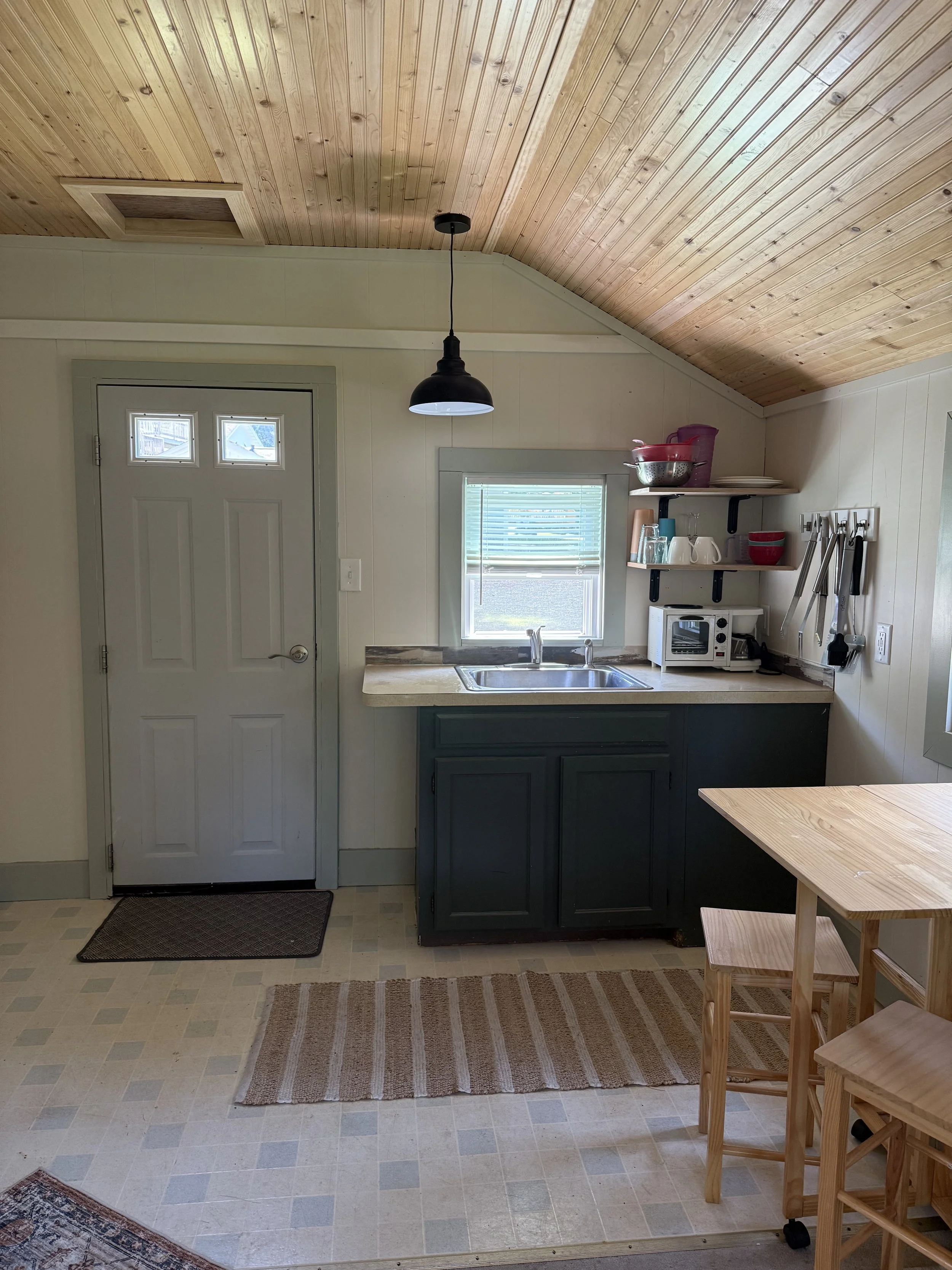 Interior of a small kitchen with wood-paneled ceiling, a window, a dark kitchen cabinet with a sink, open shelves with dishware, a microwave, and hanging utensils, a table with stools, and a door with a small window.