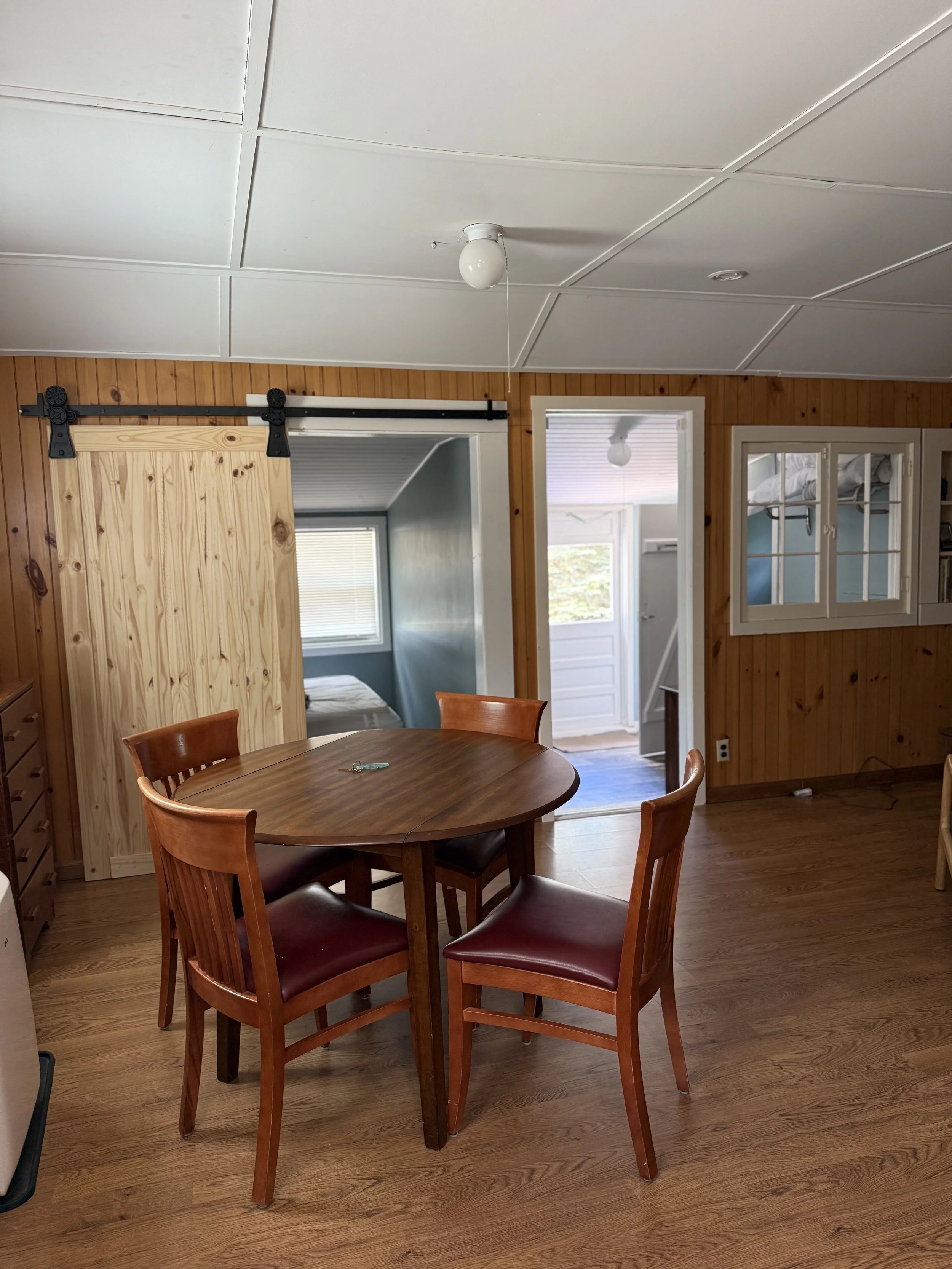 Wooden dining table with four matching chairs in a room with wood paneled walls, a sliding barn door, and an open doorway to a small sunlit room with a bed. Brookside Lake Cottages Duck Lake Rd, Grawn, MI