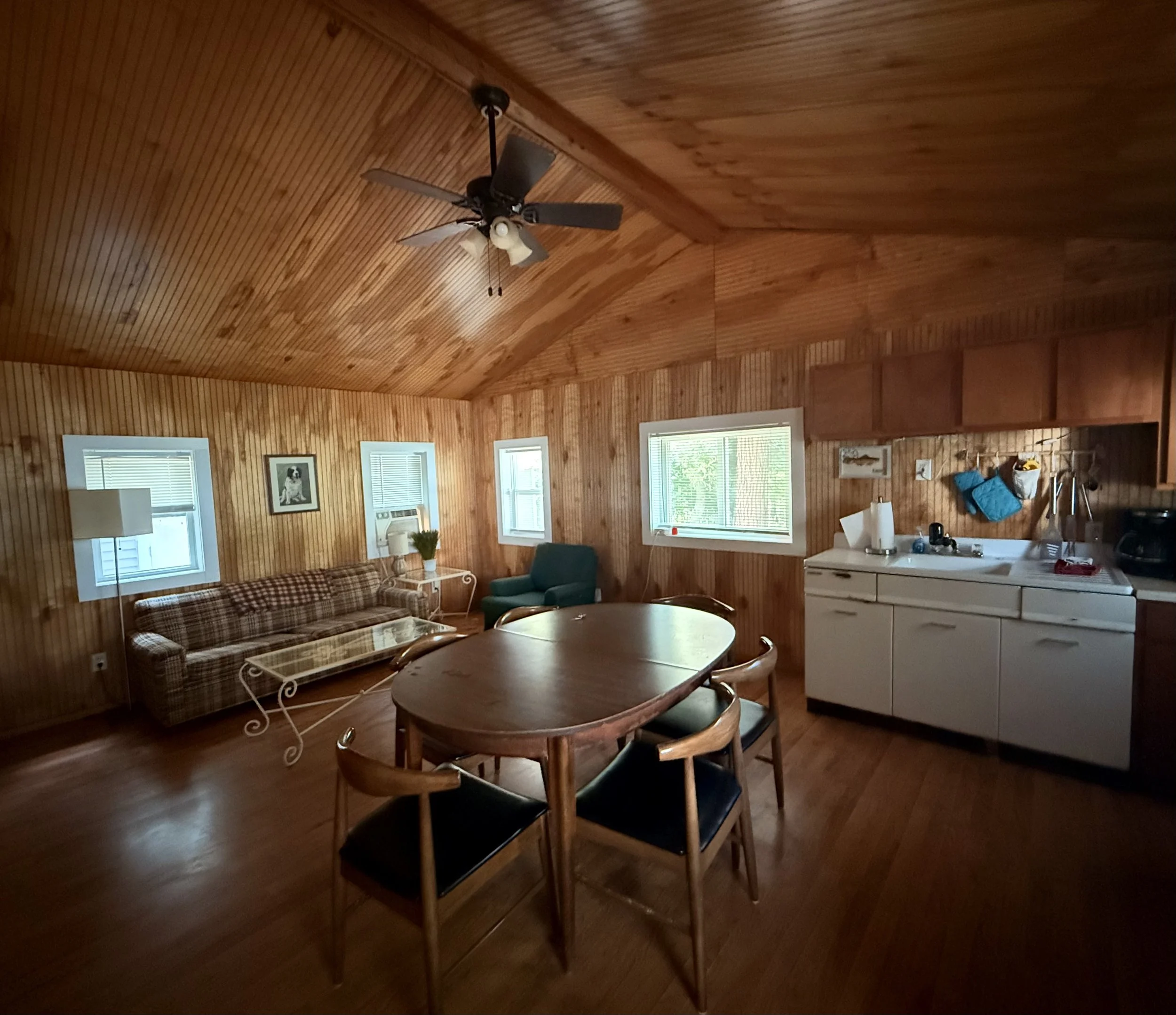 Living room with wood-paneled walls and ceiling, featuring a plaid sofa, green armchair, round wooden dining table with four chairs, white kitchen cabinets, and three windows letting in natural light. Brookside Lake Cottages Duck Lake, Grawn, MI