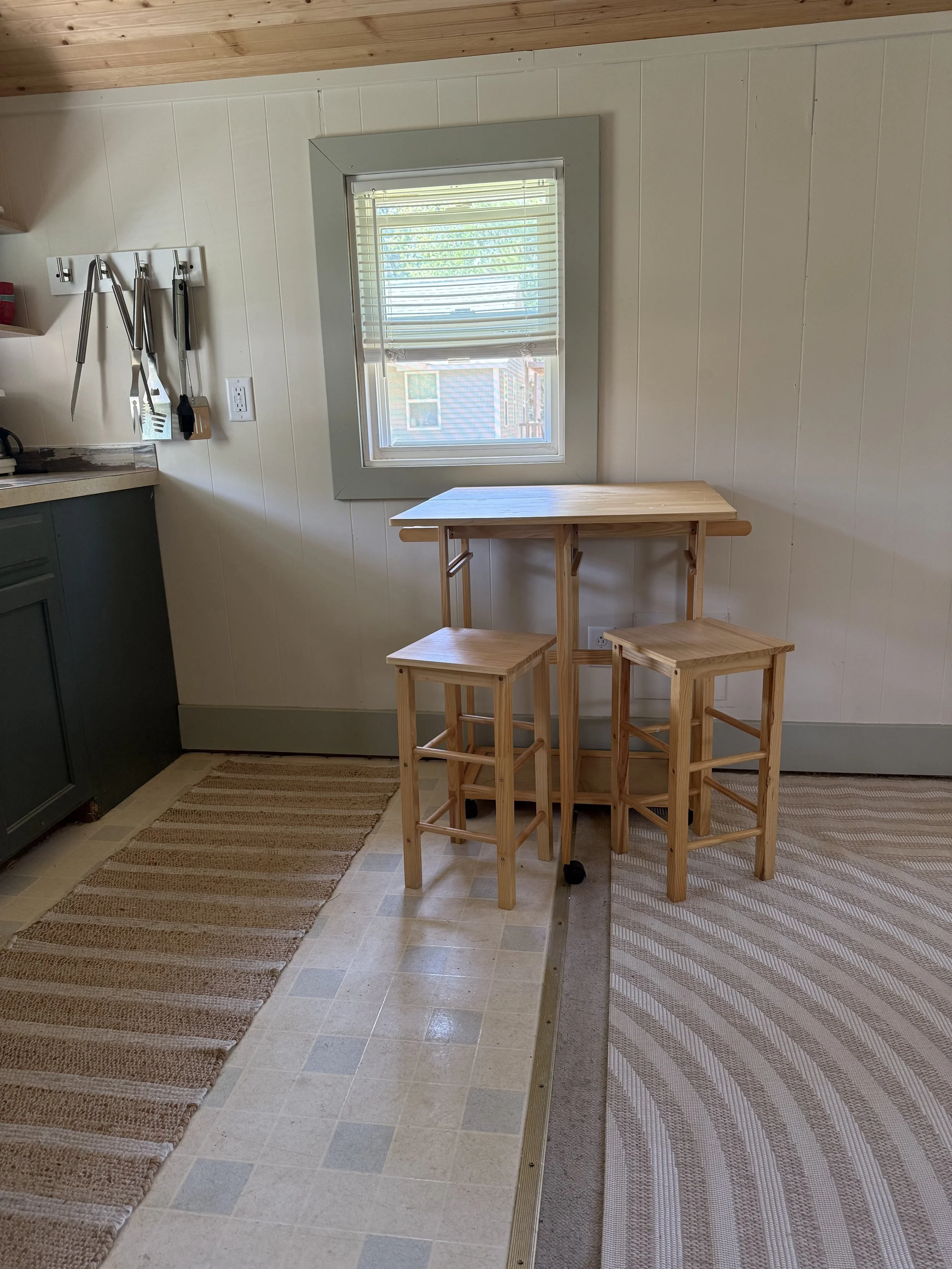 A small kitchen corner with a window, a green cabinet, and a wooden high-top table with two matching stools.