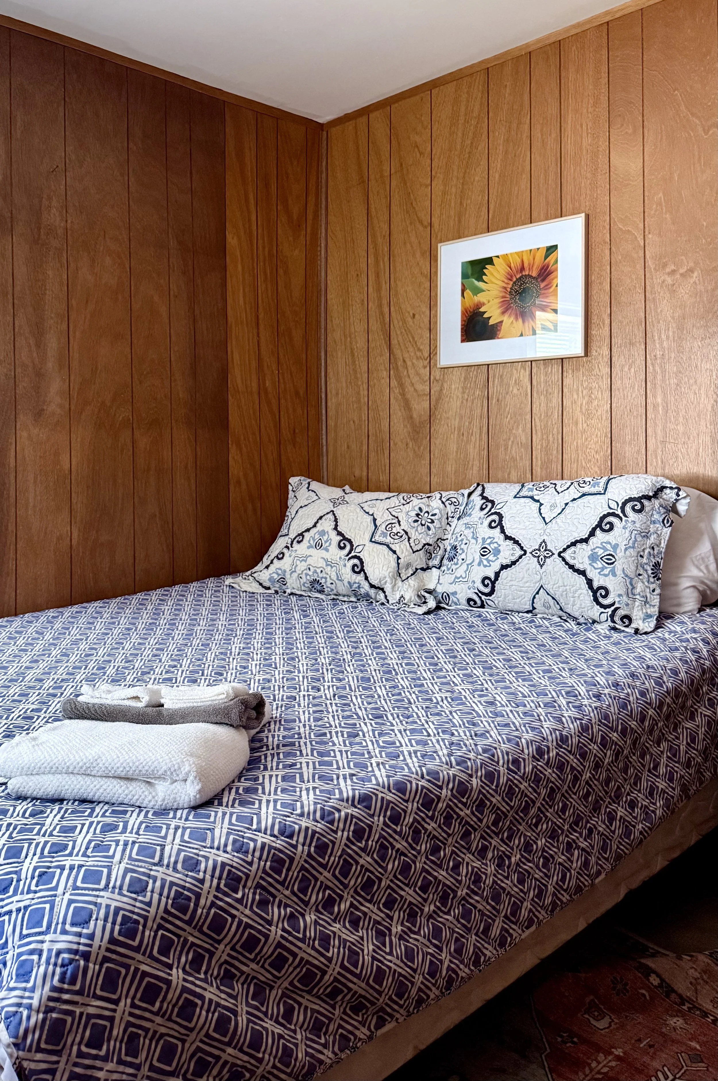 A neatly made bed with a geometric blue and white bedspread, two patterned pillows, and a folded towel, set against a wooden-paneled wall with a sunflower picture above.
