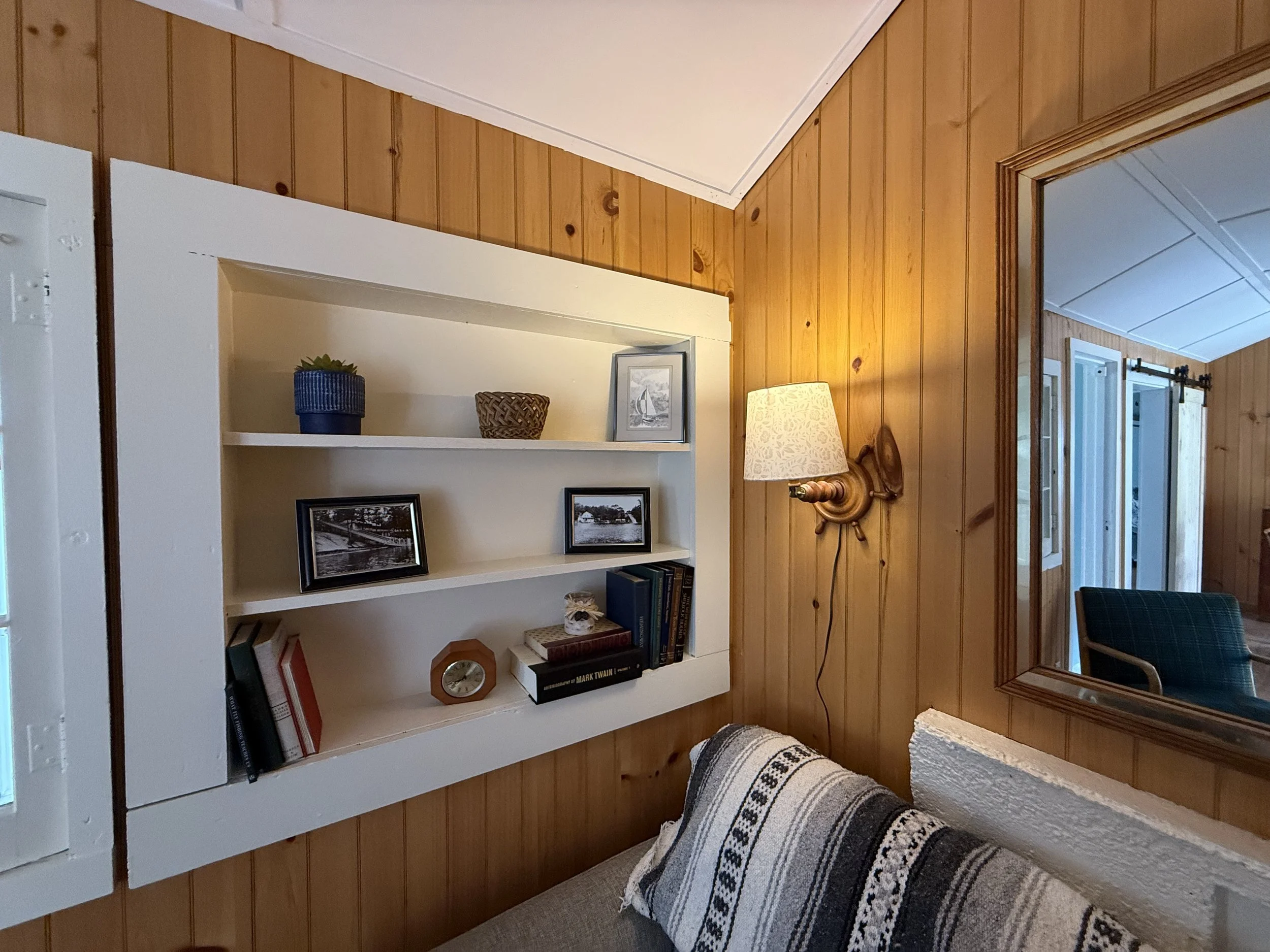 Living room corner with wood-paneled wall, wall-mounted light fixture, built-in white bookshelf with photos and books, and a mirror reflecting part of the room.